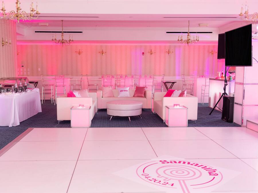 A room with a white floor, pink walls, and a chandelier. There are two white sofas, a small table, and a bar area with a TV.