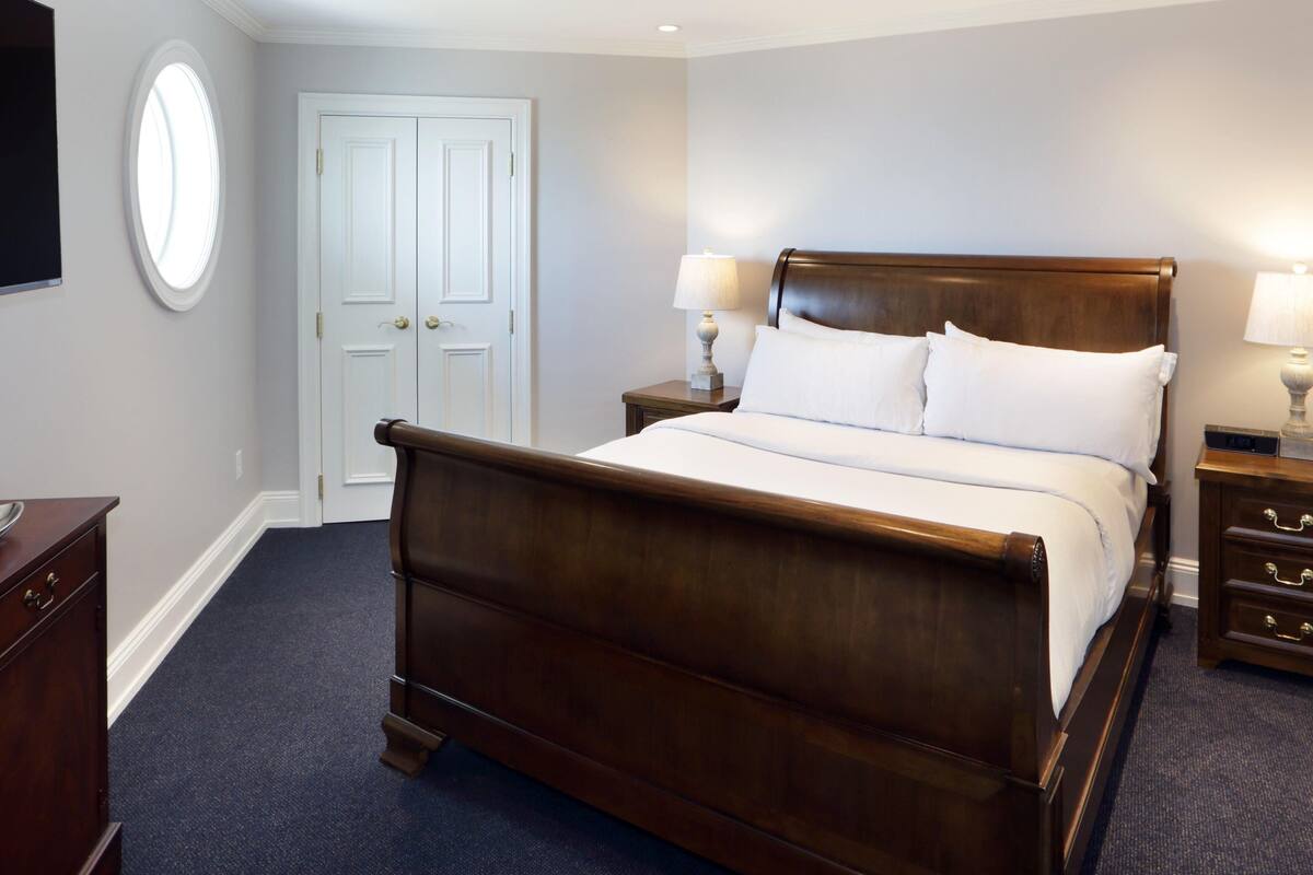 A hotel room with a wooden bed, a nightstand with a lamp, a dresser, a TV, and a door with a window.