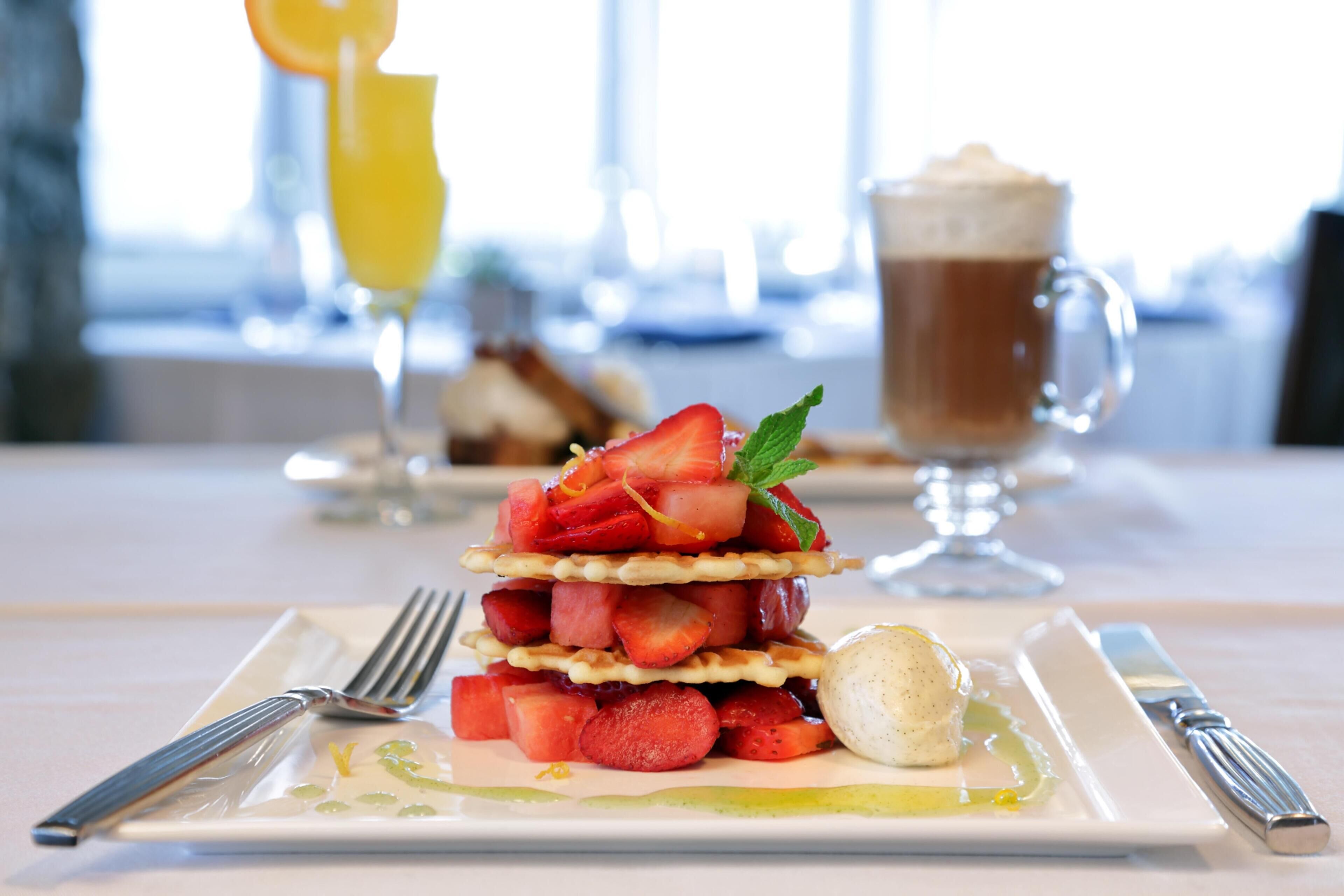 A dessert plate with a layered strawberry and waffle creation, a glass of orange juice, and a cup of coffee on a table.