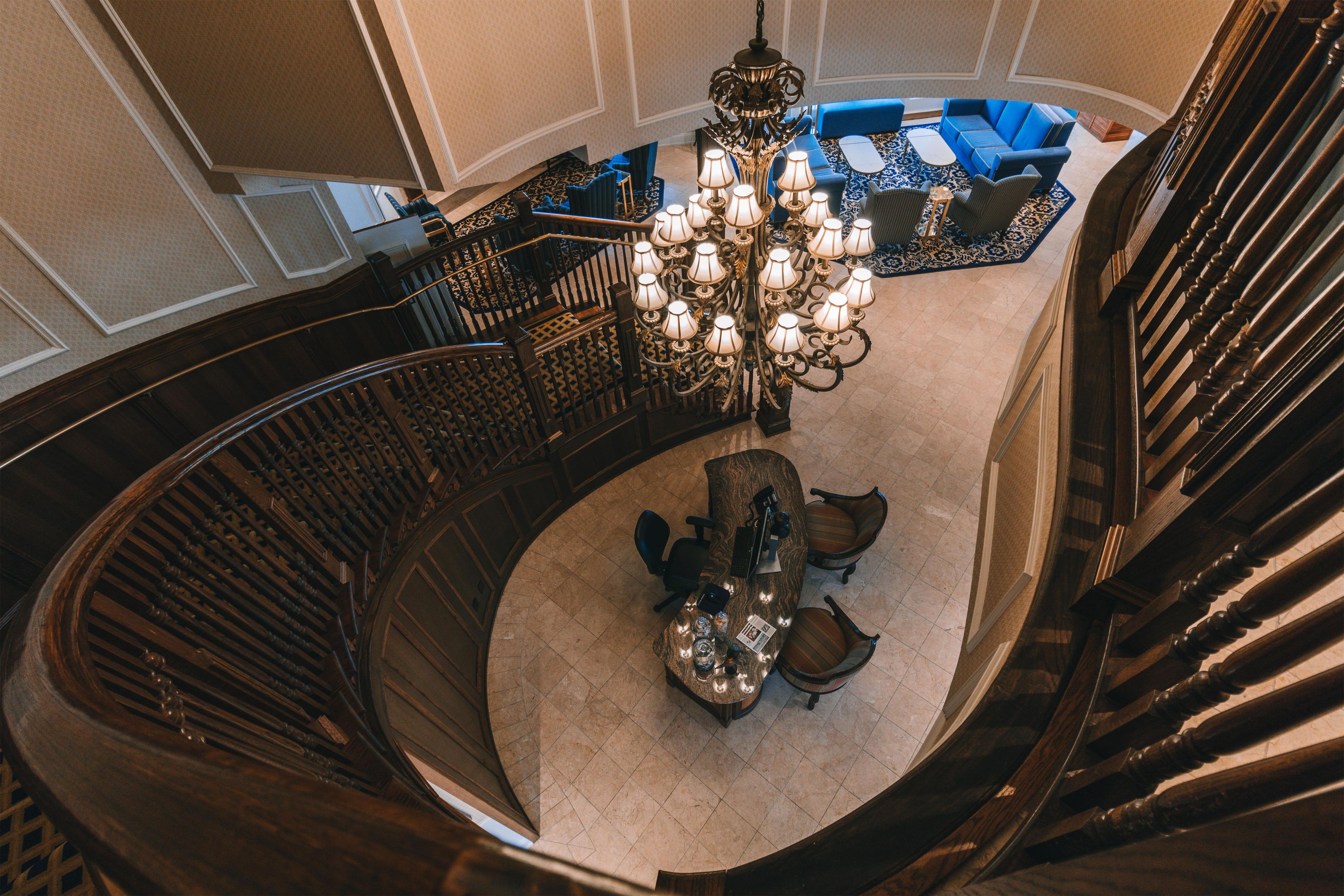 A grand staircase with a chandelier, a seating area with blue sofas, and a wooden desk with a computer.