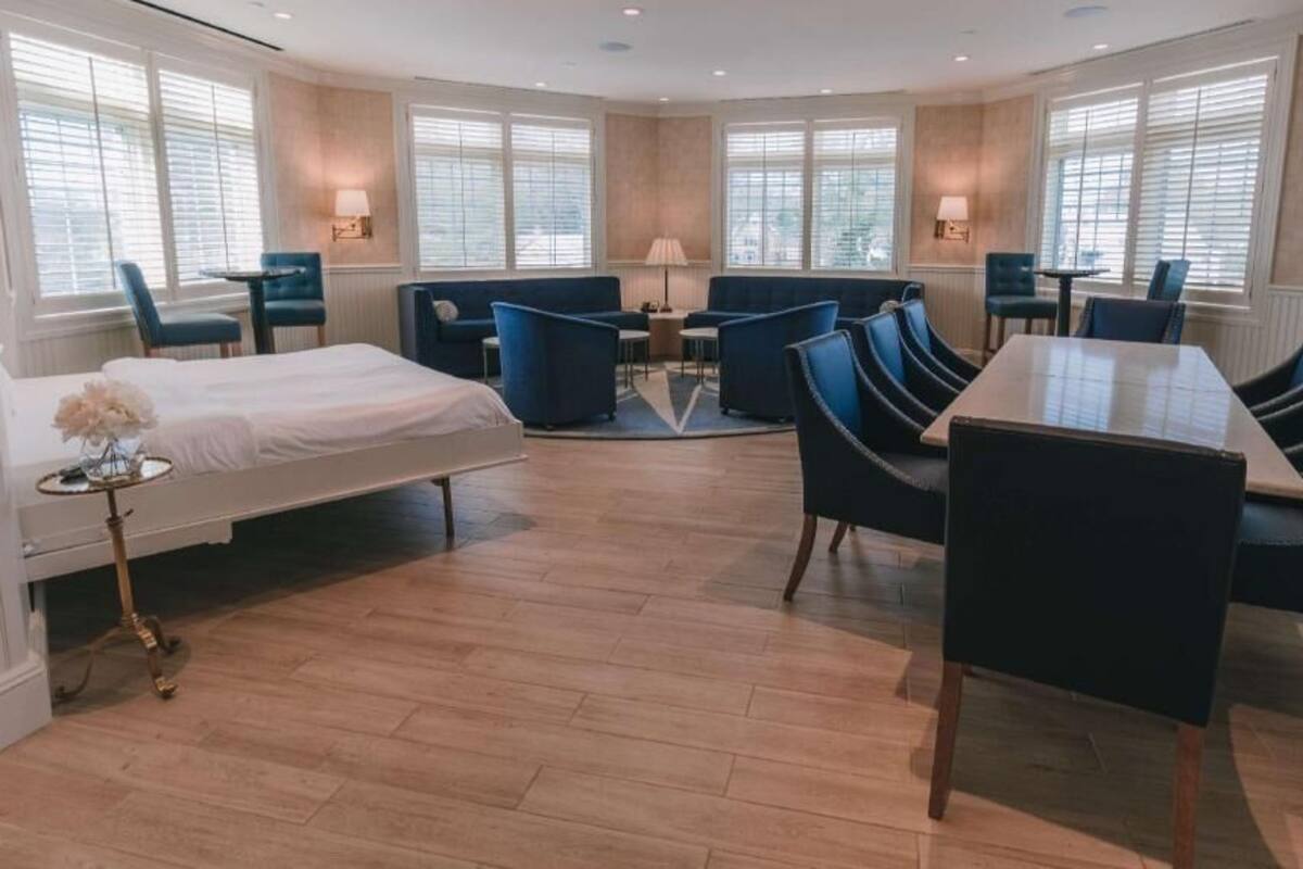 A hotel room with a bed, a dining table, multiple chairs, and large windows.