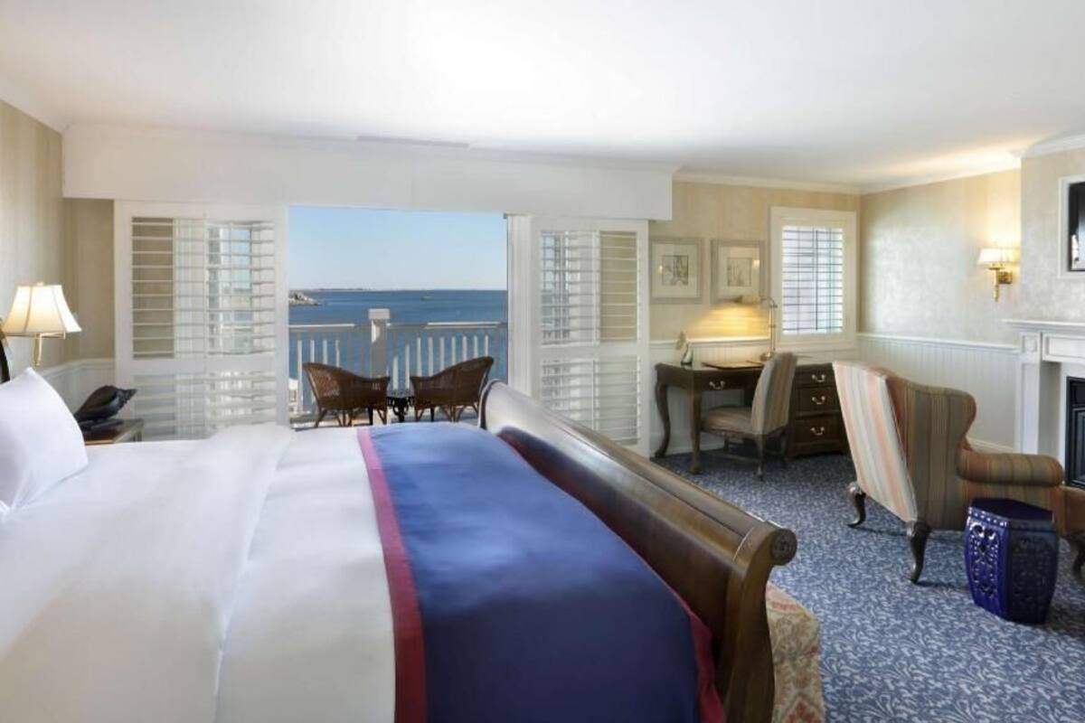 A hotel room with a bed, a desk, a chair, a fireplace, and a view of the sea.