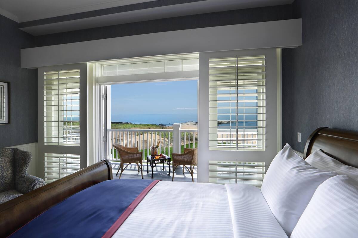 A hotel room with a large bed, two chairs, a small table, and a balcony with a view of the sea.