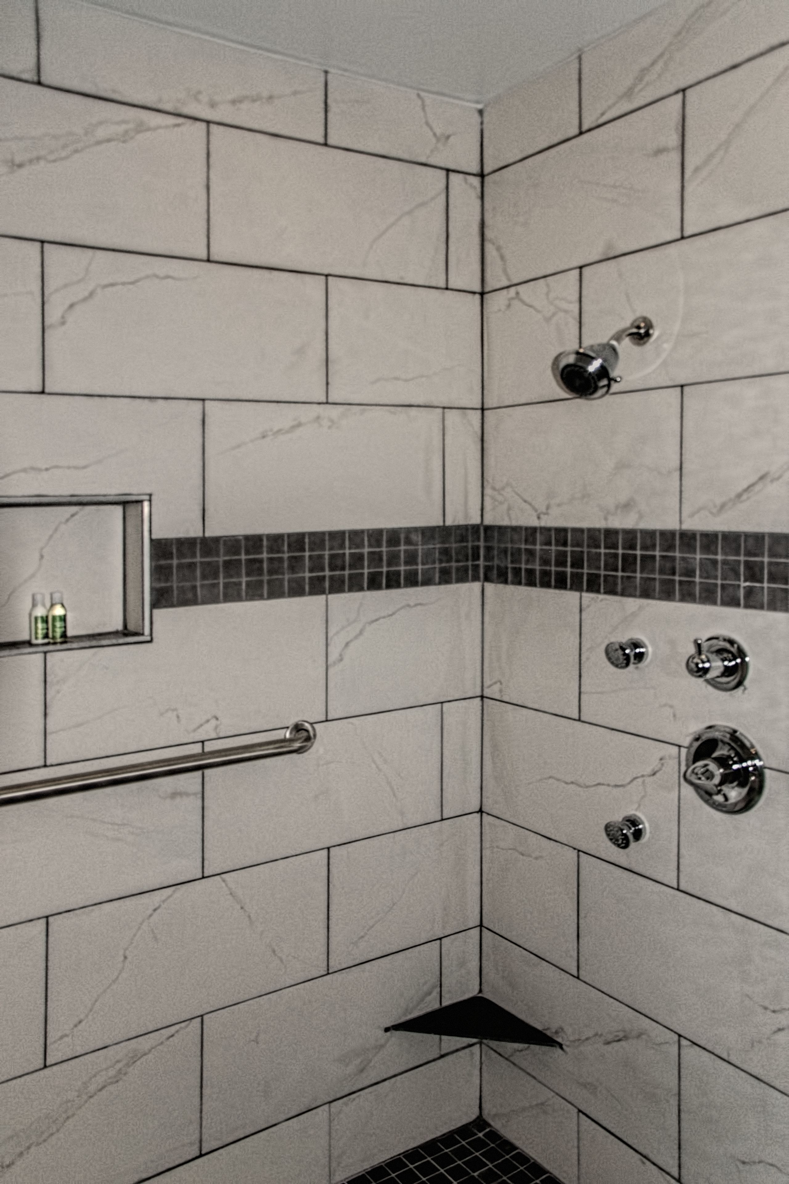 A bathroom with tiled walls, a shower with a handheld showerhead, a towel rack, and a small shelf with toiletries.