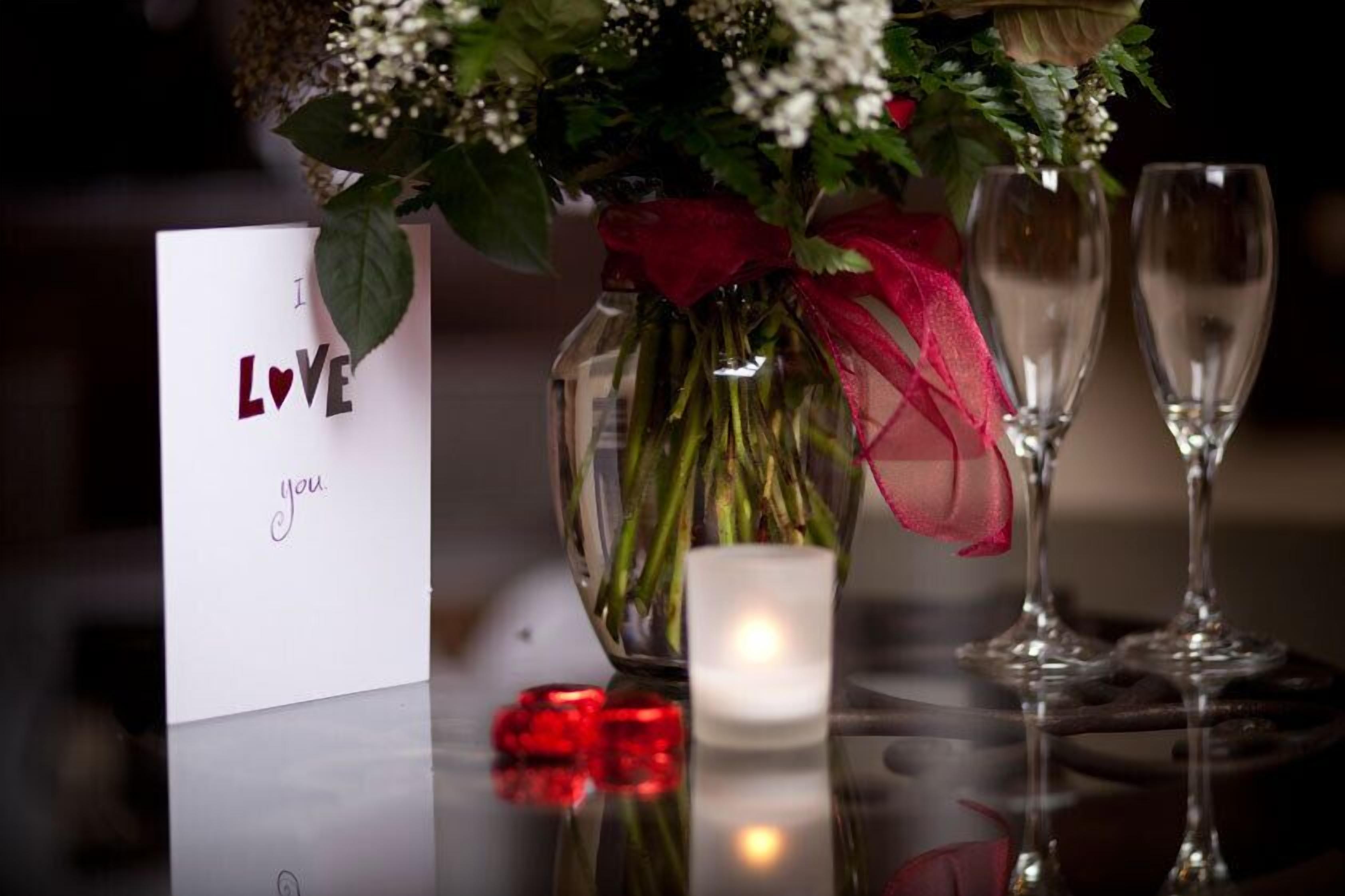 Una mesa con una tarjeta que dice “I LOVE you”, un florero con flores, una vela encendida y dos copas de vino vacías.