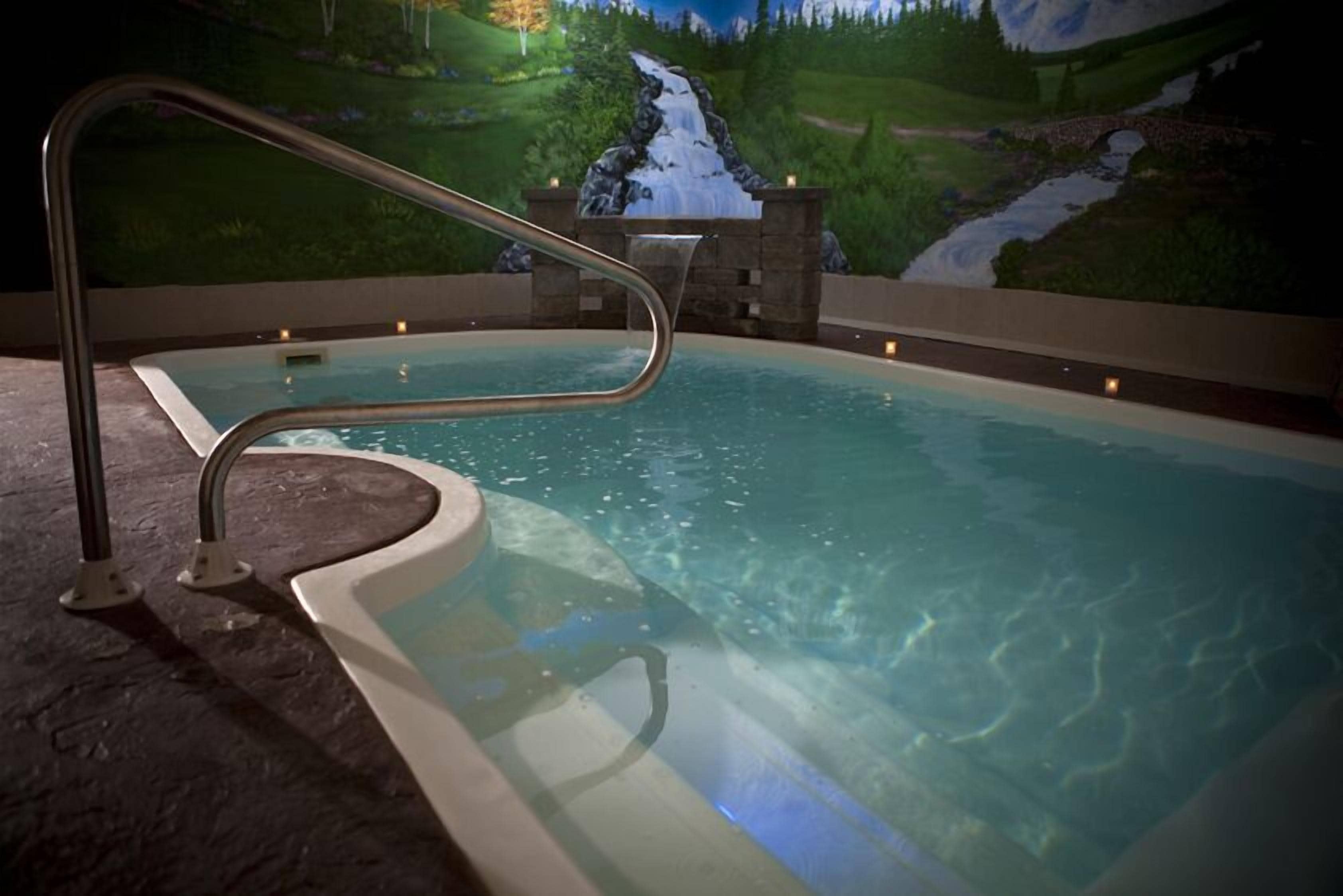 A hot tub with a waterfall mural backdrop, a metal ladder, and a stone border.