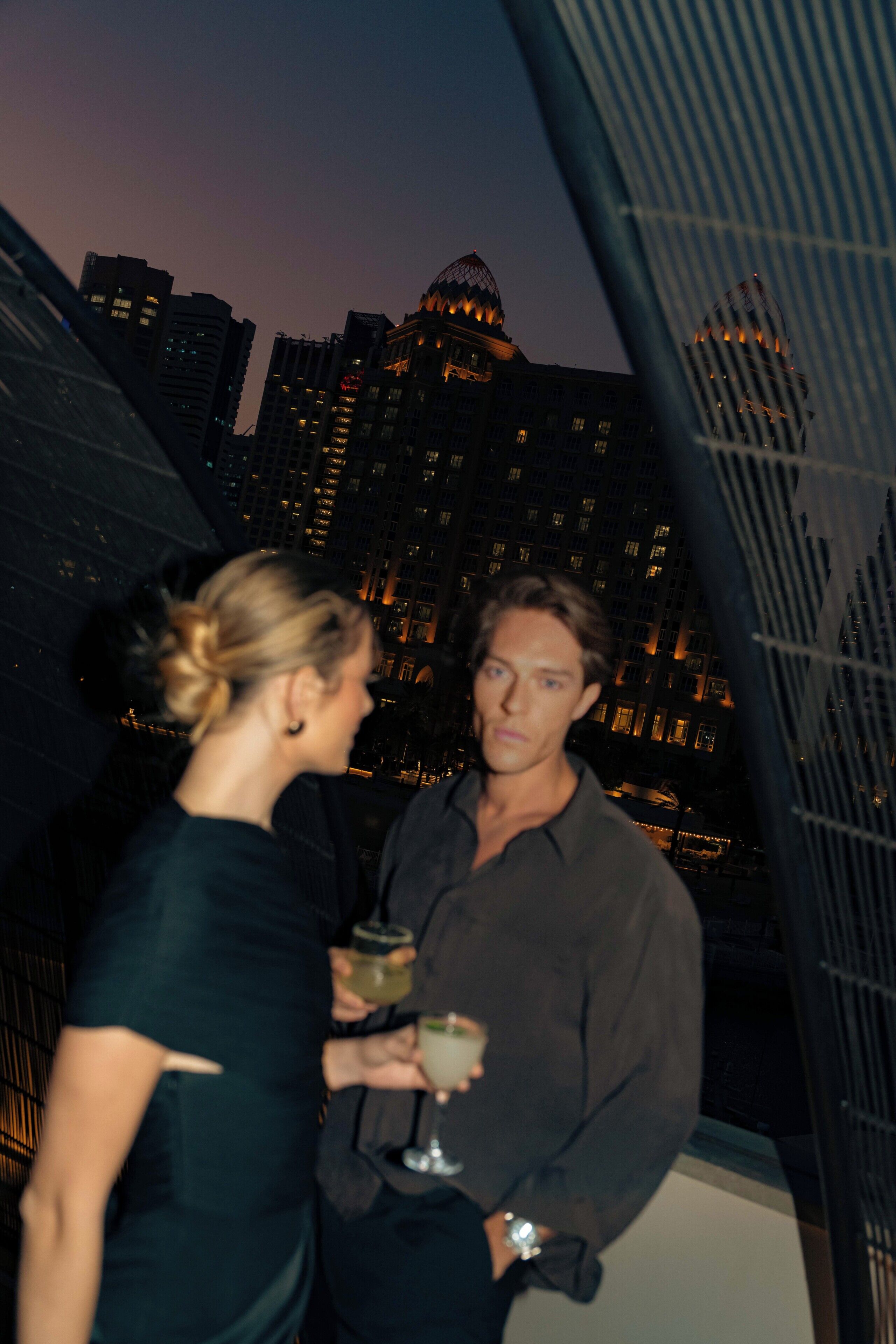 Two people standing on a balcony with a cityscape in the background.