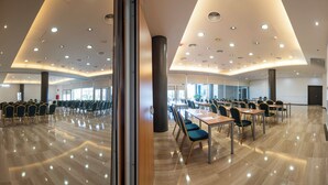 Uma sala de conferências ampla, com várias mesas e cadeiras, piso de madeira envernizada e teto moderno com iluminação embutida.