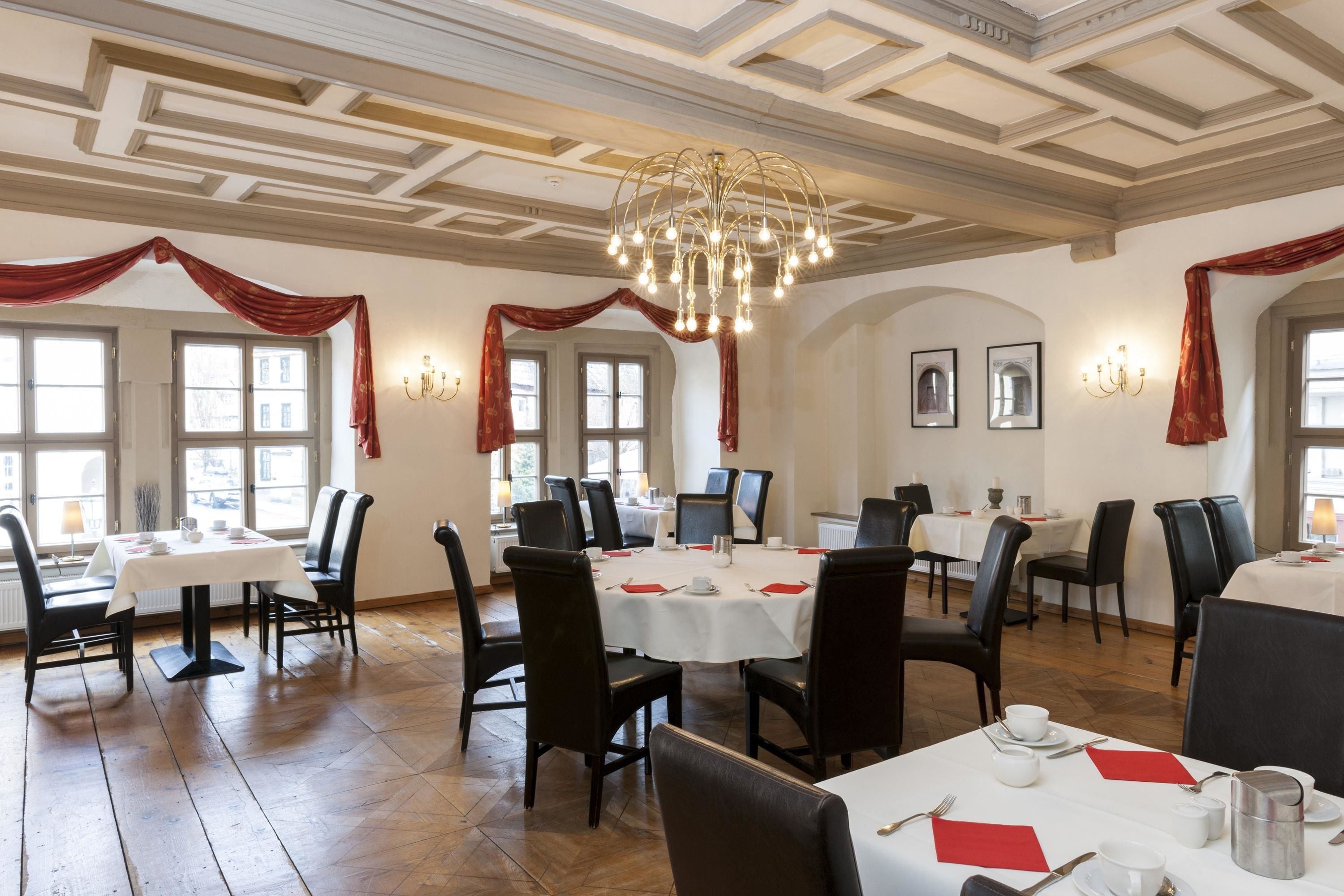 Comedor con varias mesas y sillas, una araña de techo y cortinas rojas.