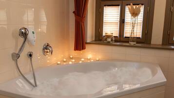 A white bathtub with a showerhead, candles, and a window with blinds.