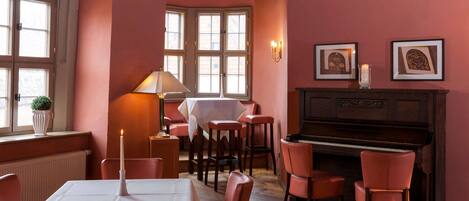 Un comedor con un piano, sillas rojas, una mesa con una vela y cuadros en la pared.