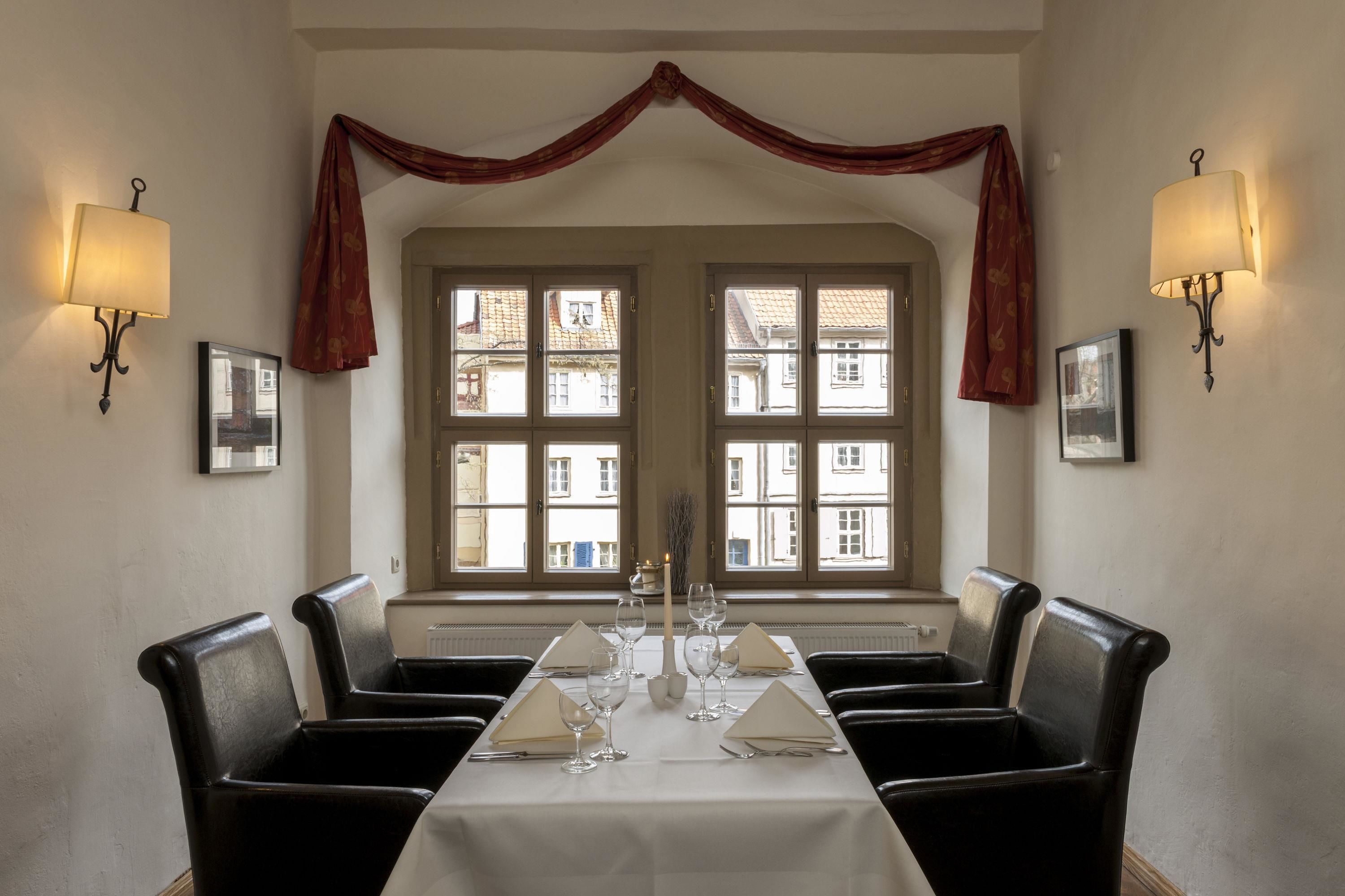 A dining room with a long table set for six, flanked by two black leather chairs, with windows and curtains in the background.
