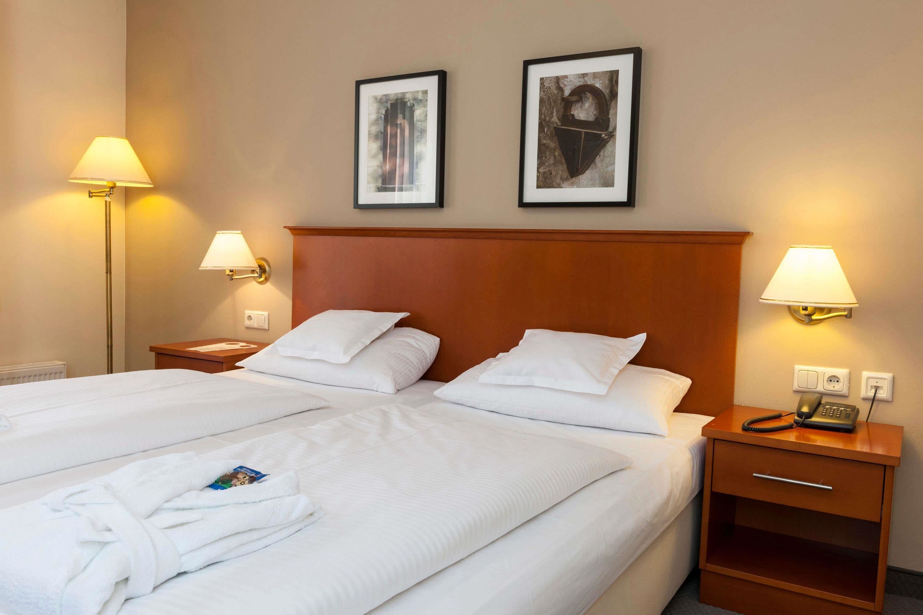 Habitación de hotel con dos camas, cabecero de madera, lámparas de noche, mesitas de noche con teléfono y dos cuadros en la pared.