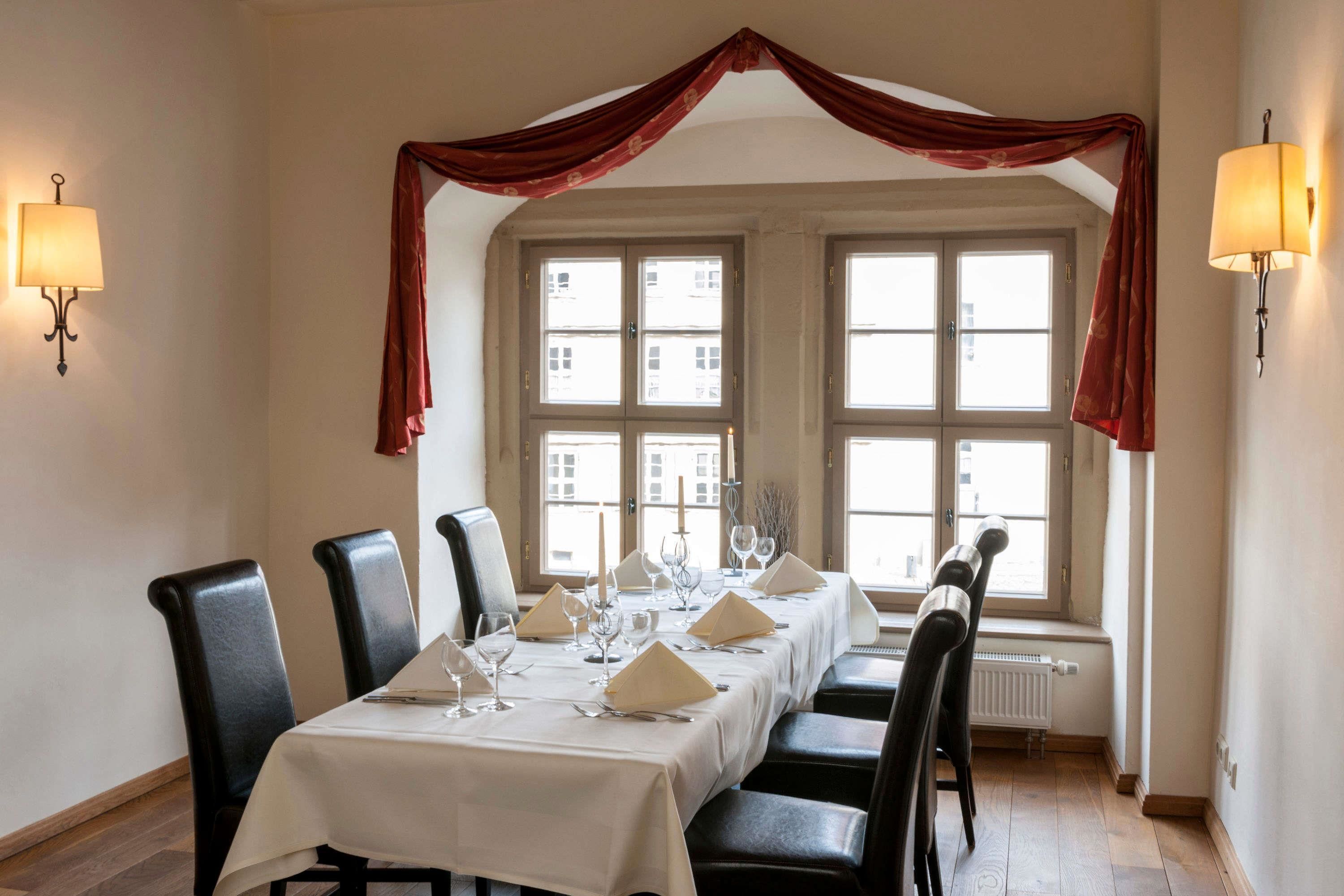 A dining area with a long table set for six, flanked by black chairs. There are two windows with curtains, and wall-mounted lamps on either side.