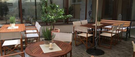 An outdoor seating area with wooden tables and chairs, potted plants, and a large umbrella providing shade.