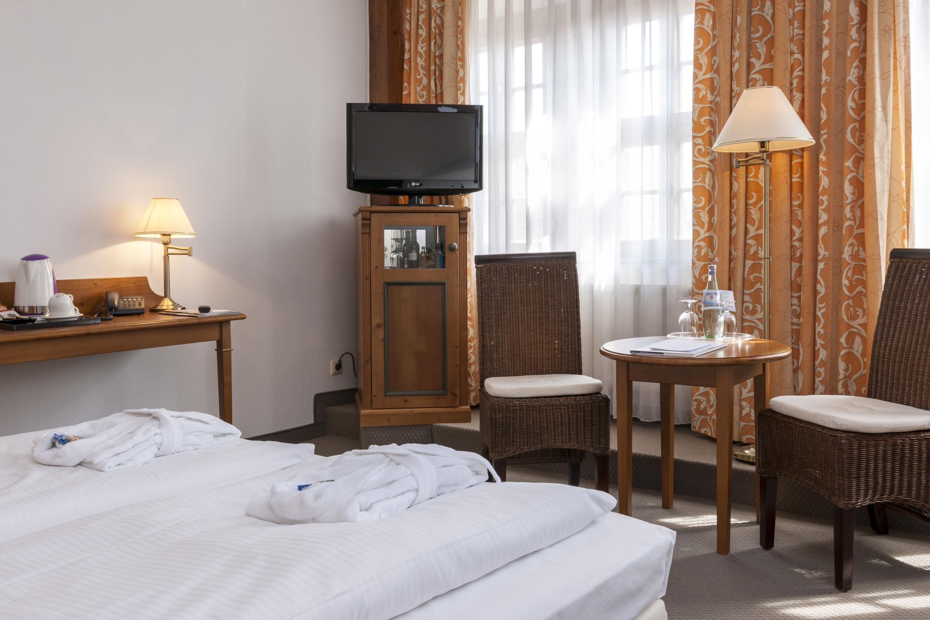 Una habitación de hotel con una cama, un televisor sobre un mueble, un escritorio con una lámpara, dos sillas de mimbre y una pequeña mesa con una lámpara.
