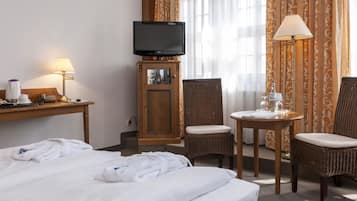 A hotel room with a bed, a television on a stand, a desk with a lamp, two wicker chairs, and a small table with a lamp.