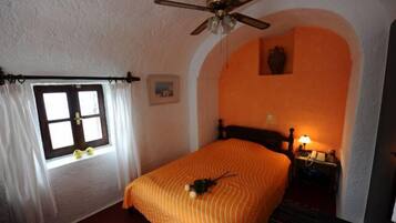 A bedroom with a bed, a ceiling fan, a nightstand with a lamp, a window with curtains, and an arched wall.