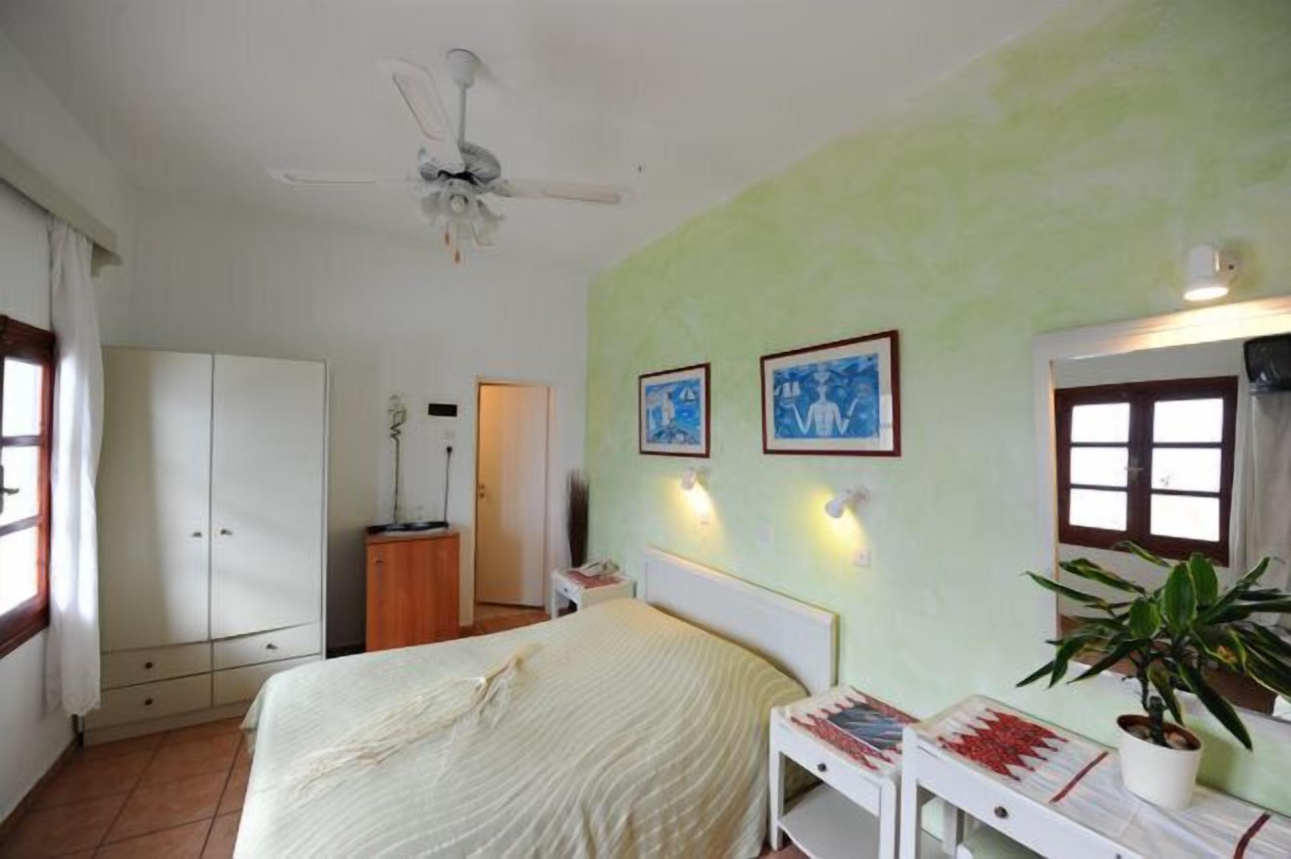 A bedroom with a bed, a ceiling fan, a wardrobe, a bedside table with a lamp, and two framed pictures on the wall.