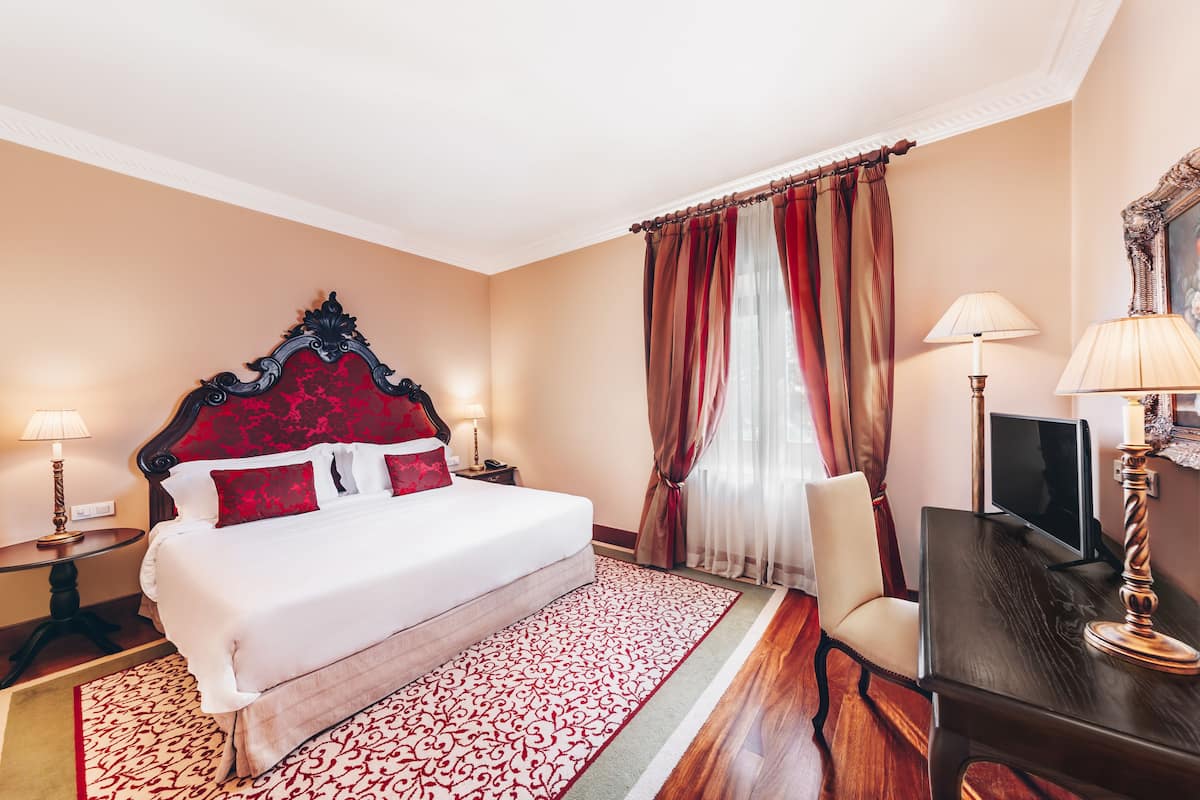 A hotel room with a large bed, a desk, a chair, a TV, and a lamp.