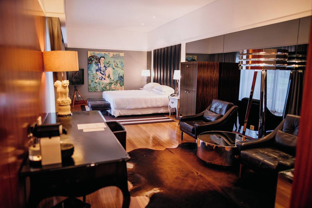 A hotel room with a bed, a desk, a chair, a lamp, and a painting on the wall.