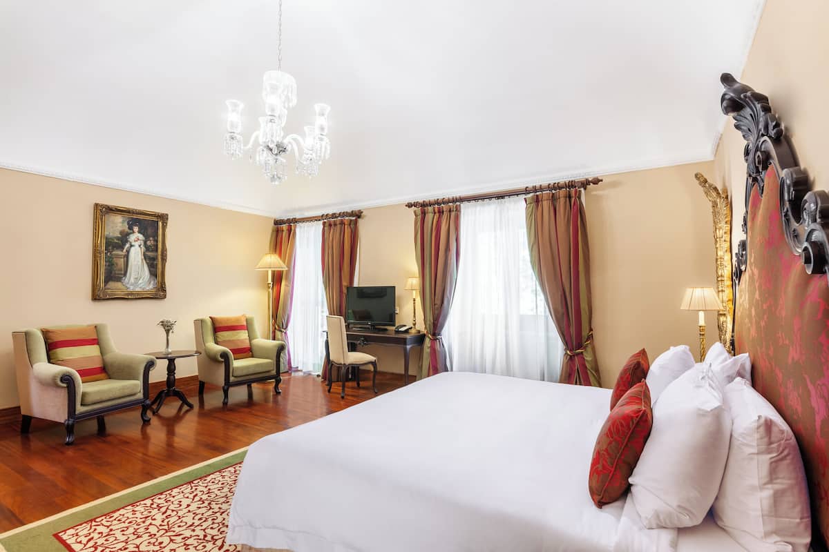A hotel room with a large bed, two armchairs, a desk with a computer, a television, and a chandelier.