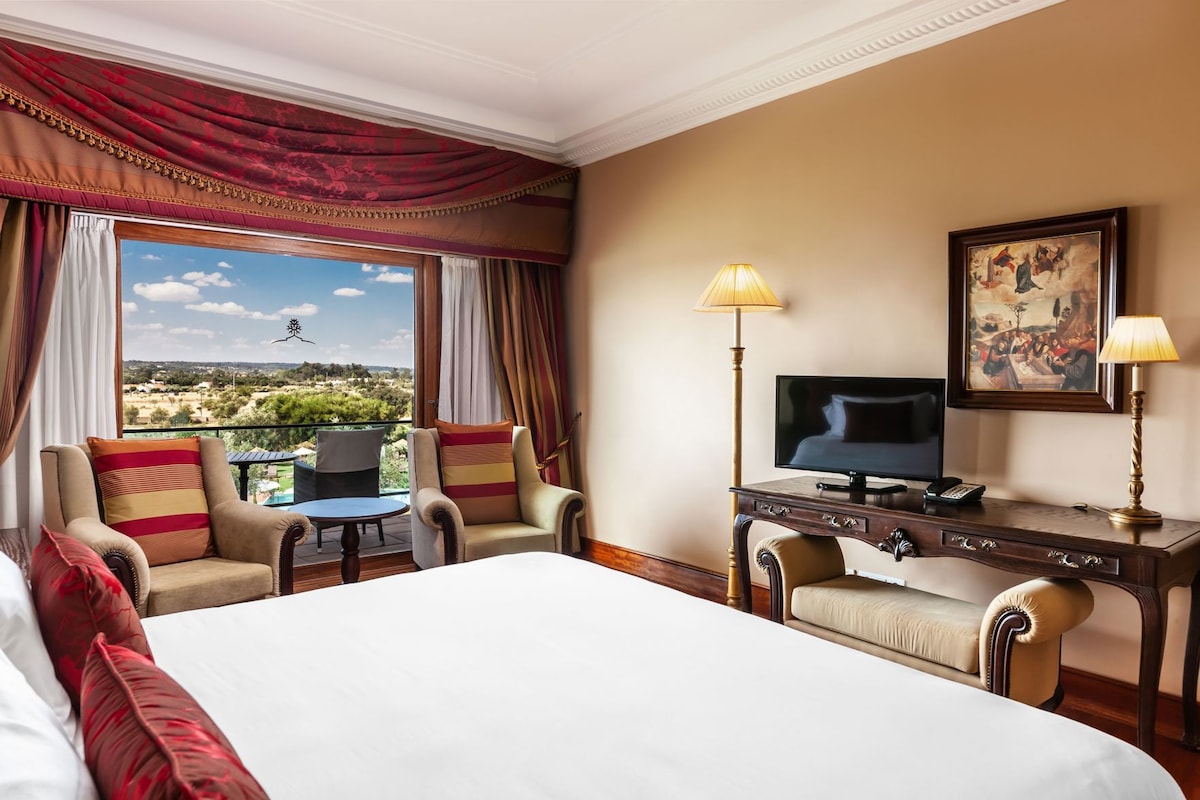 A hotel room with a large bed, two armchairs, a small table, a desk with a TV, and a painting on the wall.