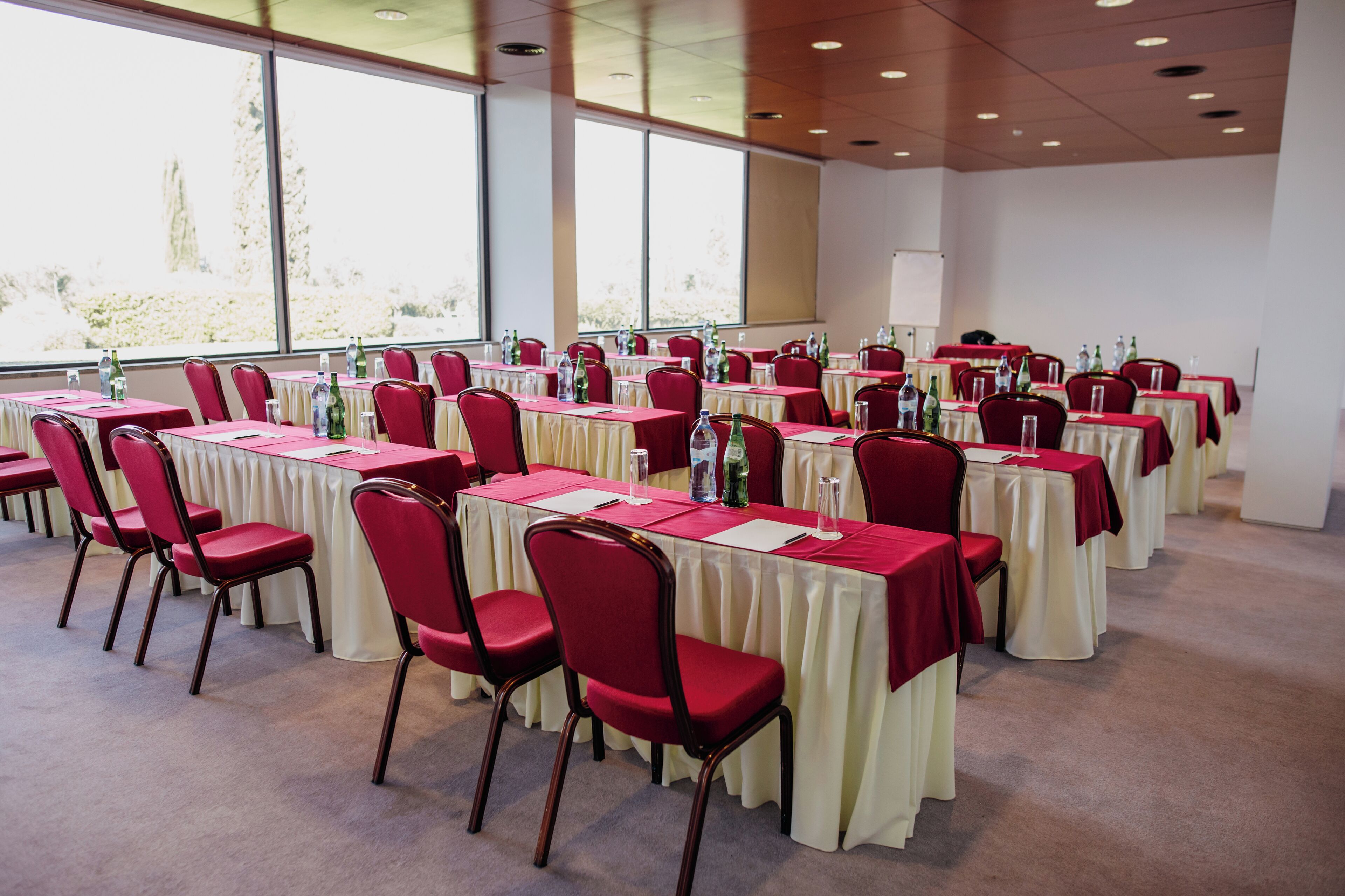 Un salón de conferencias con filas de mesas y sillas, amplias ventanas y suelo alfombrado.