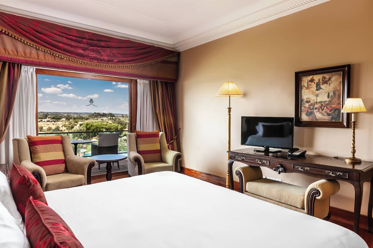 A hotel room with a large bed, two armchairs, a small table, a desk with a TV, and a balcony with a view.