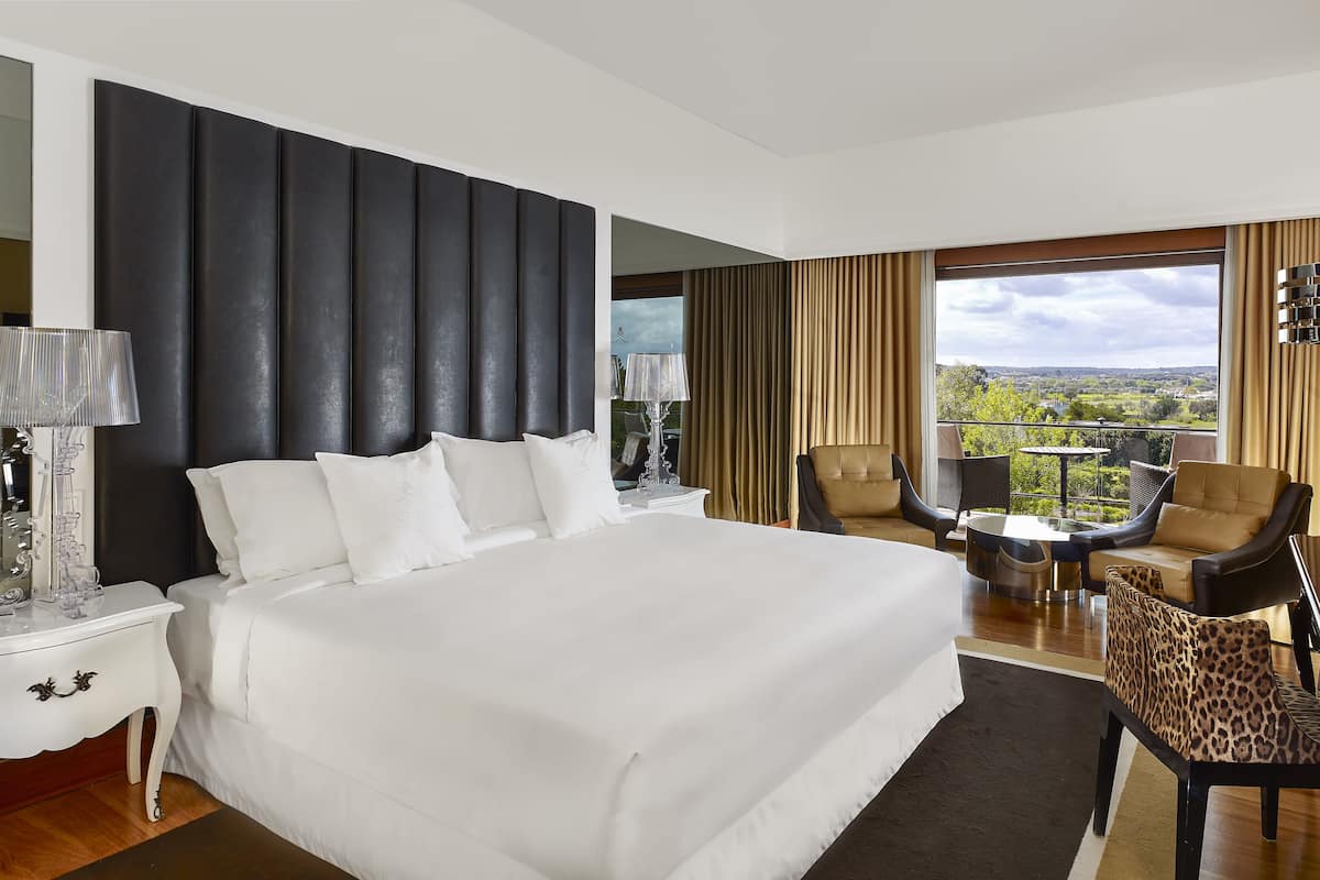 A hotel room with a large bed, a desk with a lamp, a chair, a small table, and a view of the outdoors.