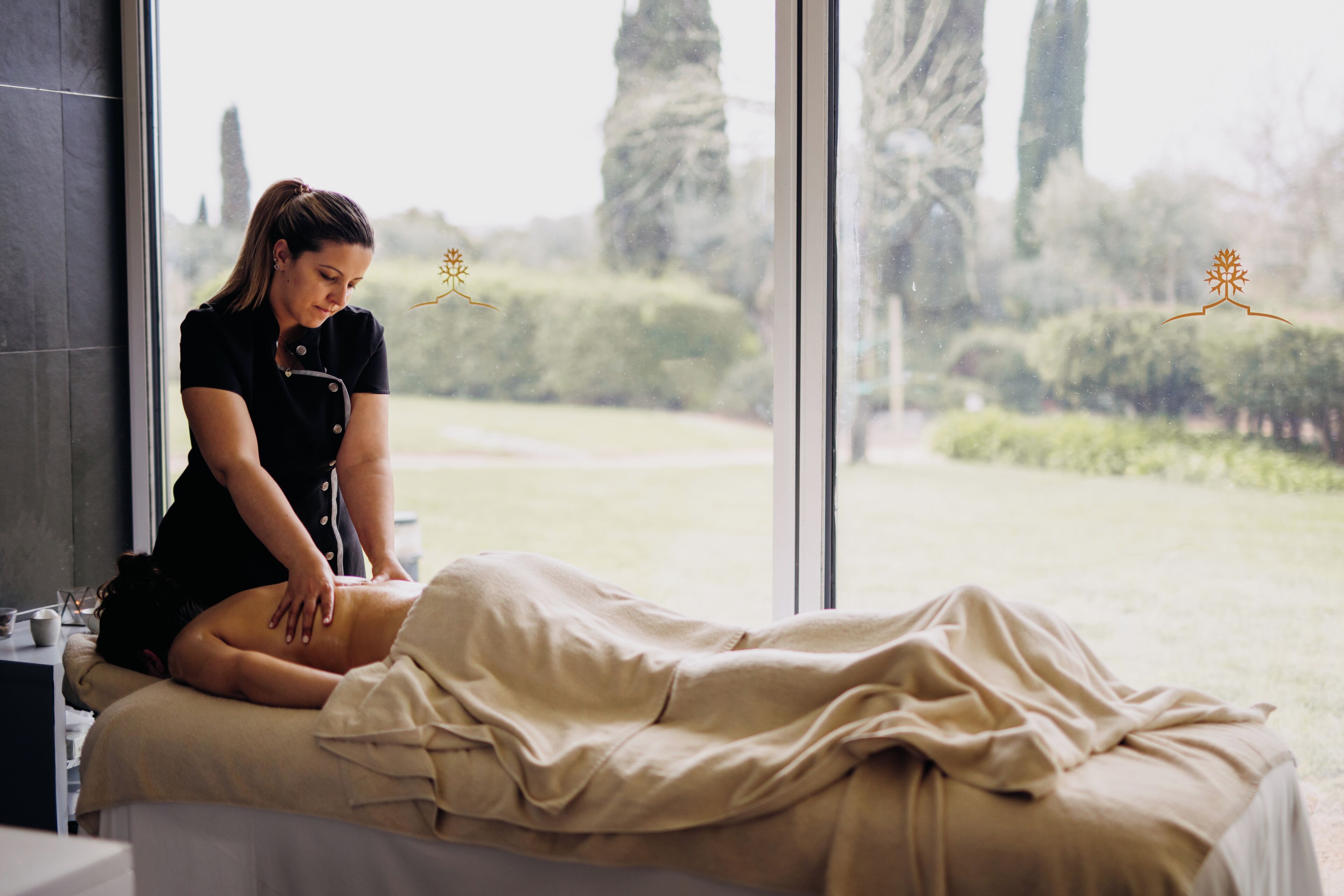 A massage therapy room with a large window, a massage table, and a therapist.