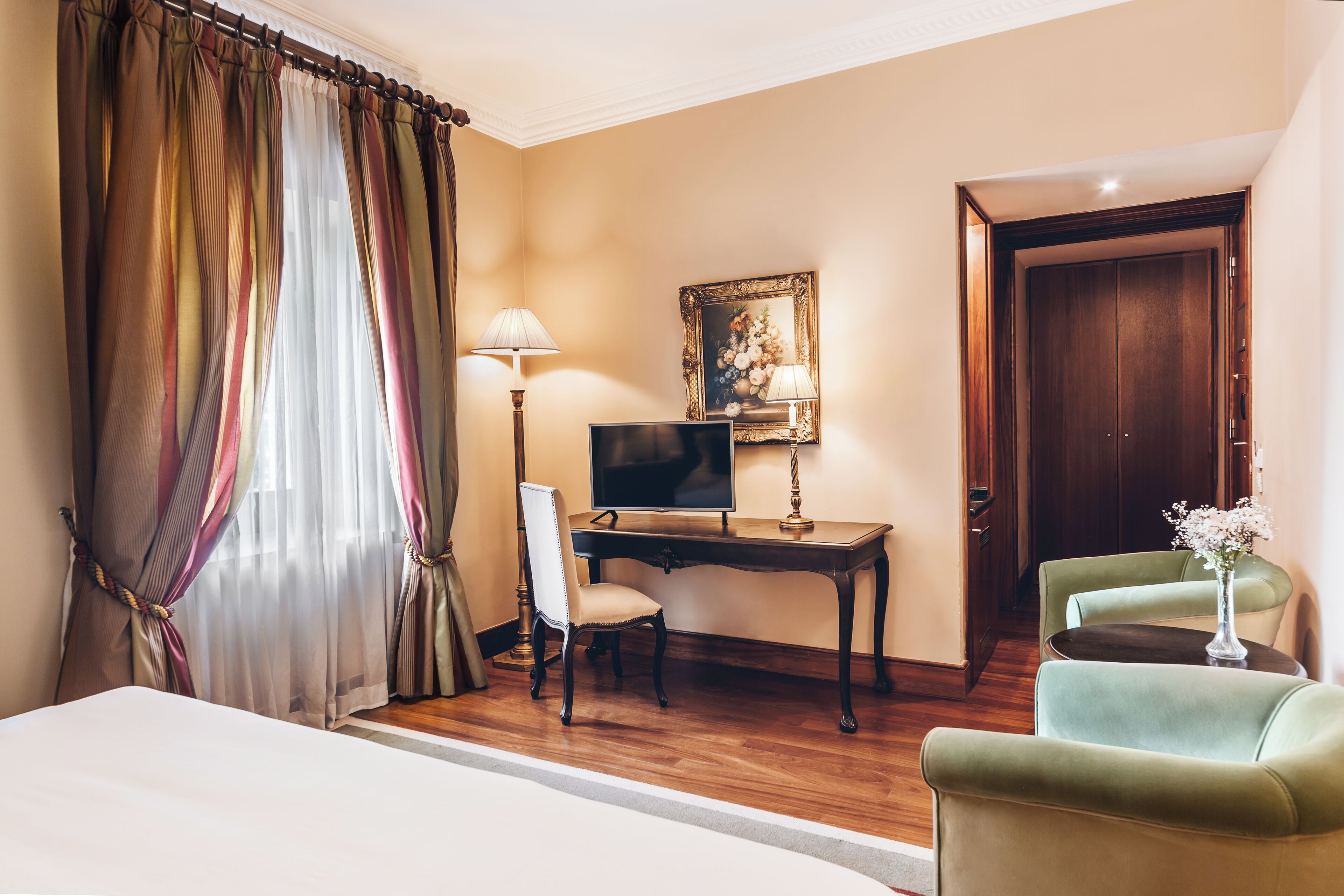 A hotel room with a bed, desk, computer, two chairs, a lamp, and a painting on the wall.