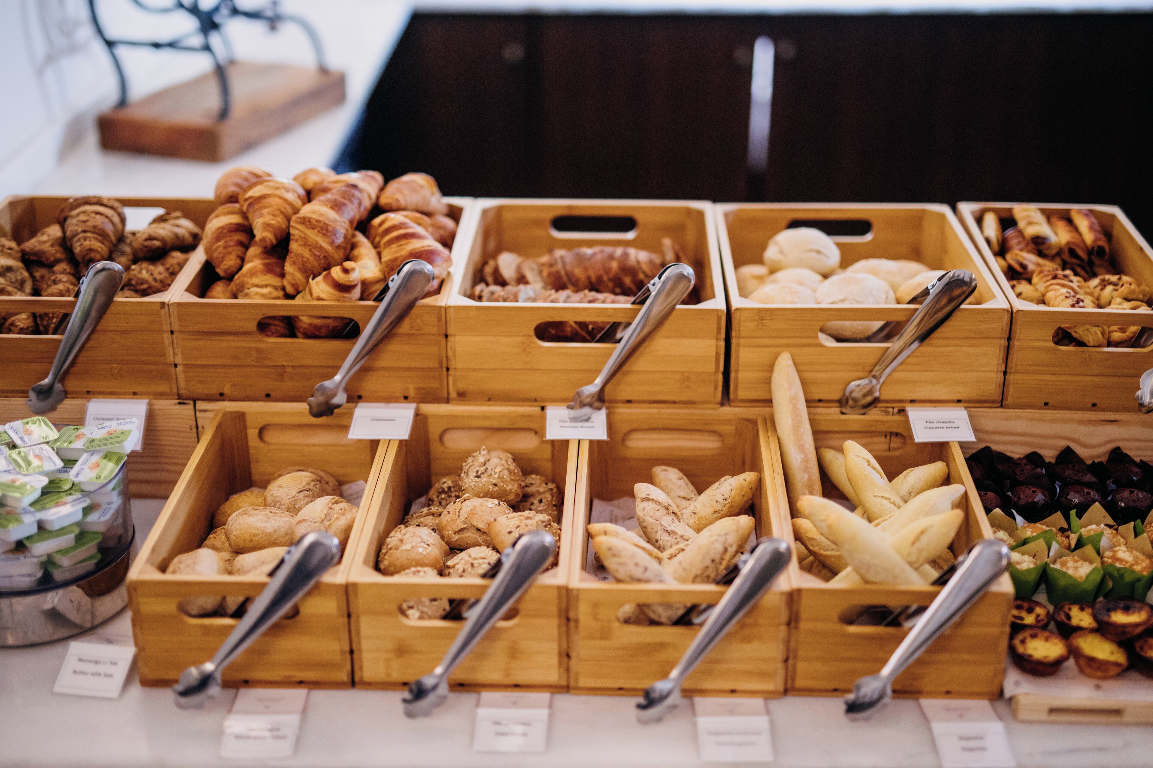 A variety of baked goods displayed in wooden crates with metal tongs, each labeled with a price tag.