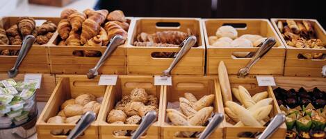 A variety of baked goods displayed in wooden crates with metal tongs, each labeled with a price tag.