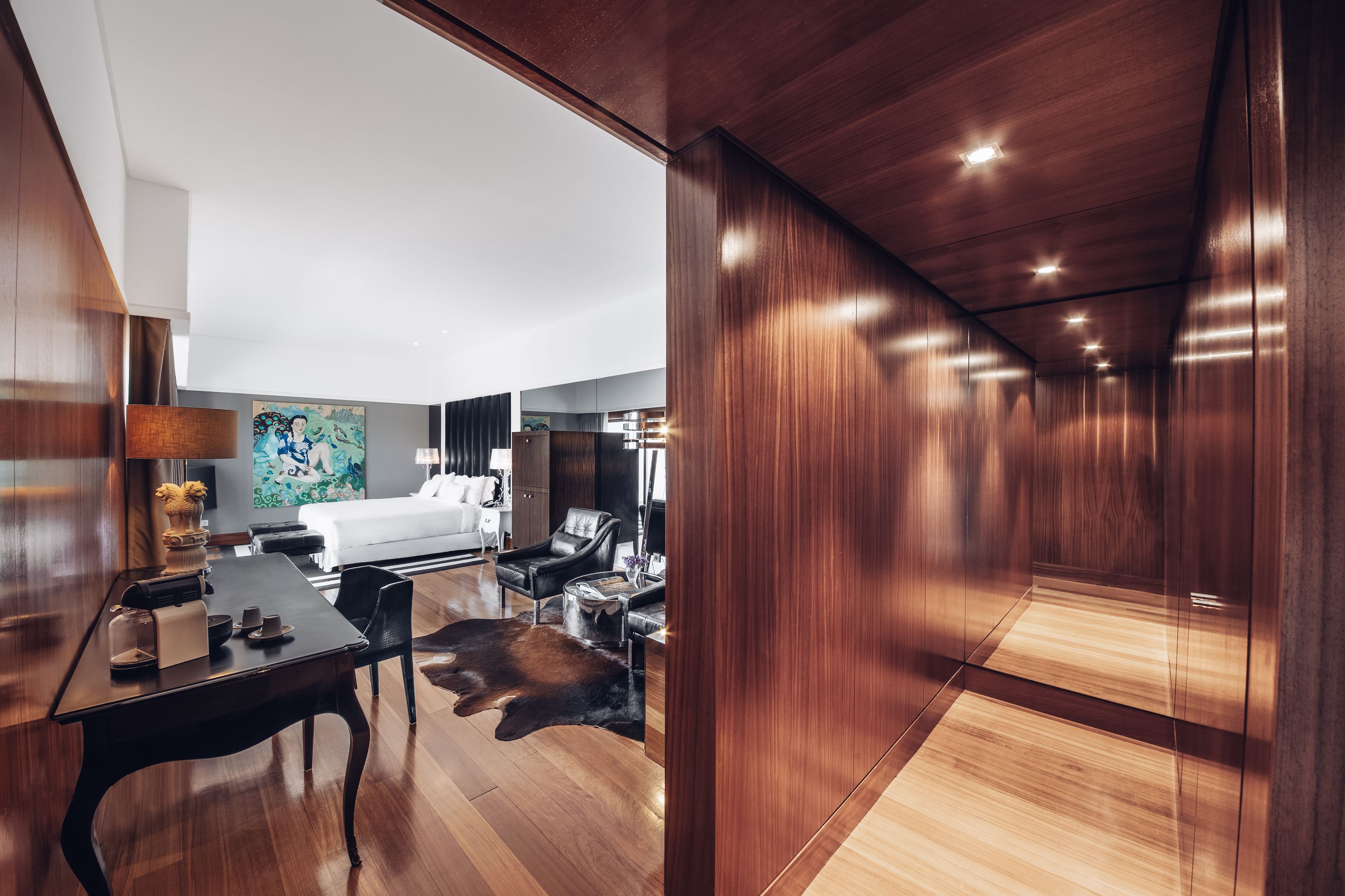 A modern hotel room with a wooden floor, a dining area with a table and chairs, a bed with a white headboard, and a large abstract painting on the wall.