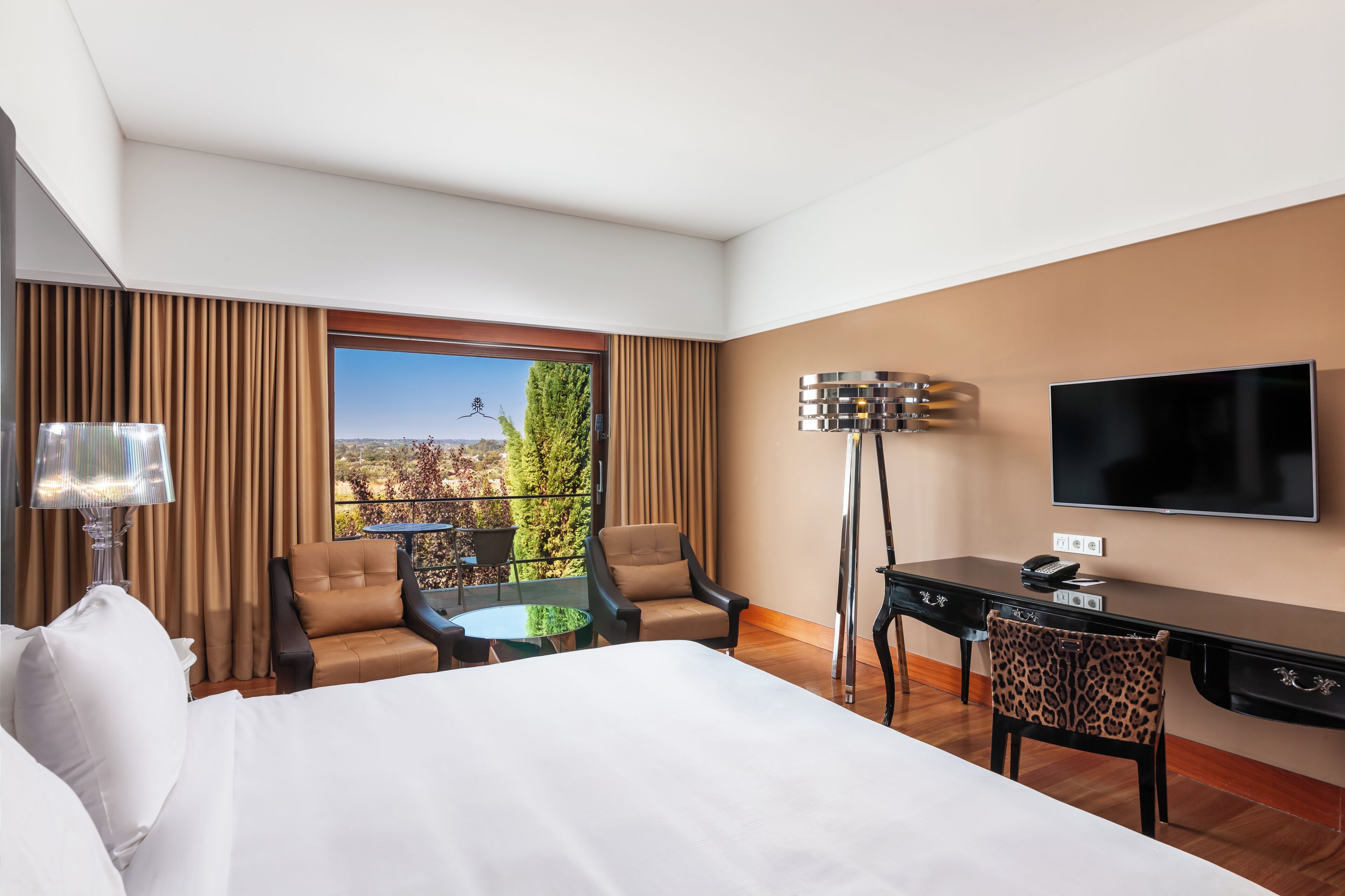 A hotel room with a bed, two armchairs, a desk, a television, and a view of the outdoors.