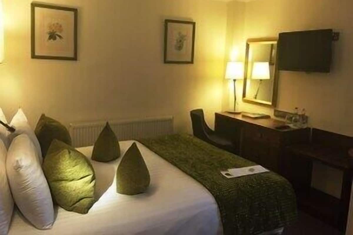A hotel room with a bed, pillows, a desk, a chair, a lamp, and a television on the wall.