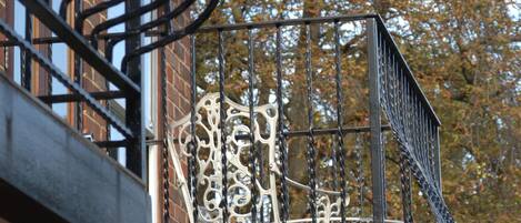 A wrought iron balcony railing with intricate designs, a brick building, and autumn foliage in the background.