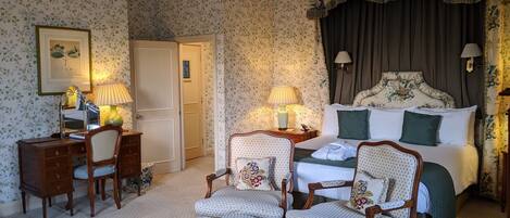Habitación de hotel con papel tapiz floral, una cama con almohadas verdes, un escritorio con lámpara y dos sillones.