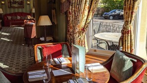 A dining area with a round table set for two, a red sofa, and a window with floral curtains. - Cotswold Lodge Hotel (Oxford)