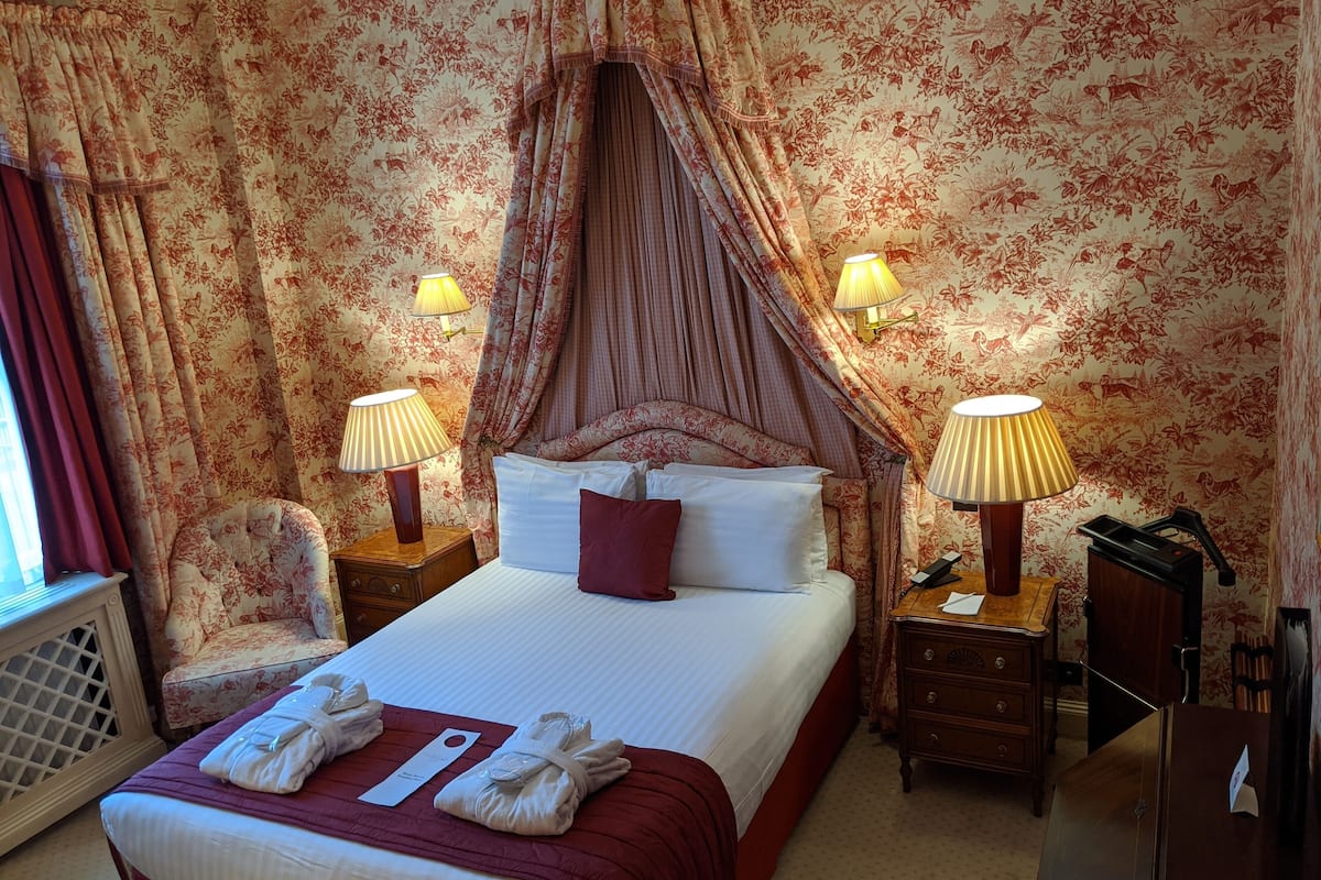 A hotel room with a canopy bed, floral wallpaper, two bedside lamps, a wooden nightstand, a chair, and a desk with a lamp.