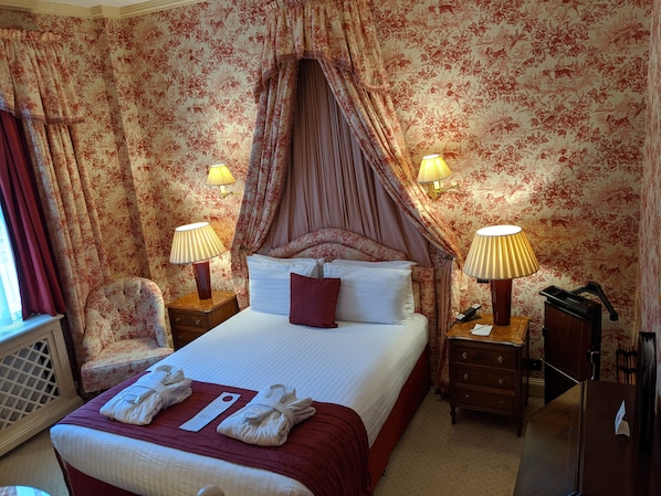 Una habitación de hotel con cama a dosel, papel tapiz floral, dos lámparas de noche, una mesita de noche de madera, una silla y un escritorio con lámpara.