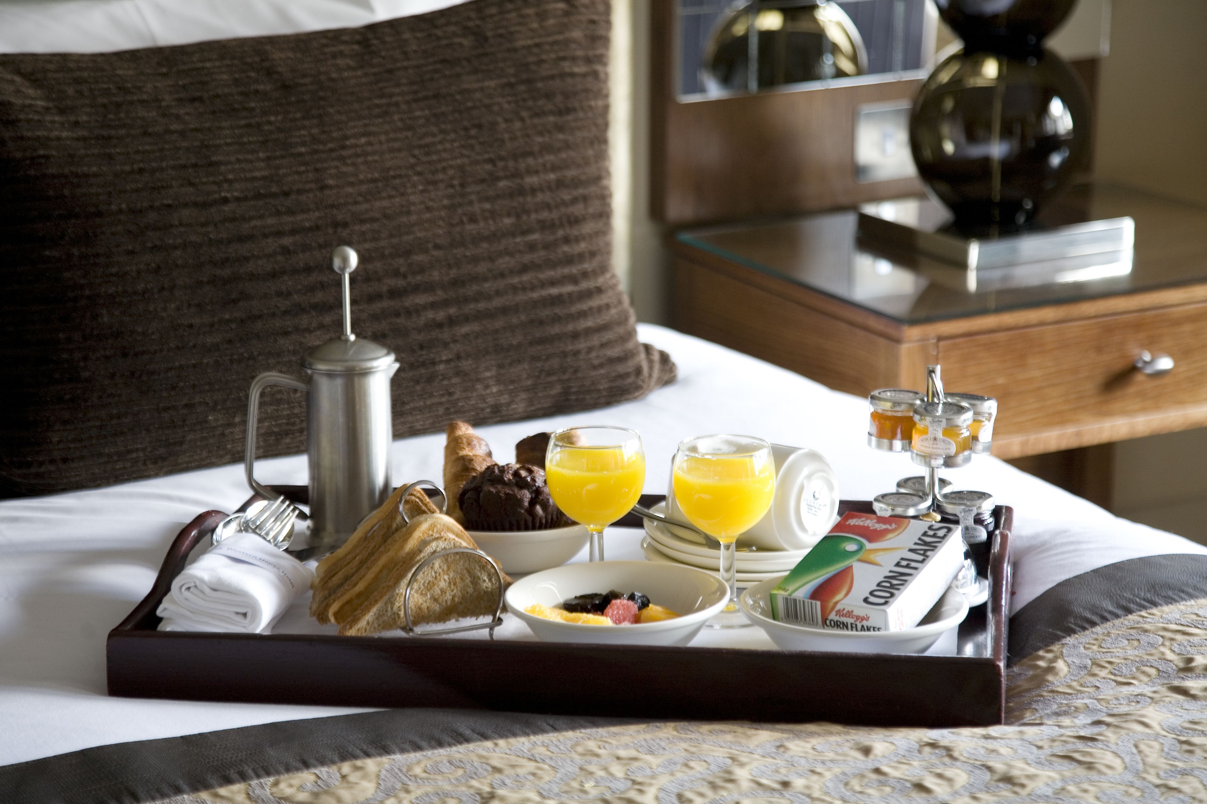 A breakfast tray on a bed with croissants, muffins, orange juice, cereal, and a coffee pot.