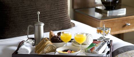 A breakfast tray on a bed with croissants, muffins, orange juice, cereal, and a coffee pot.
