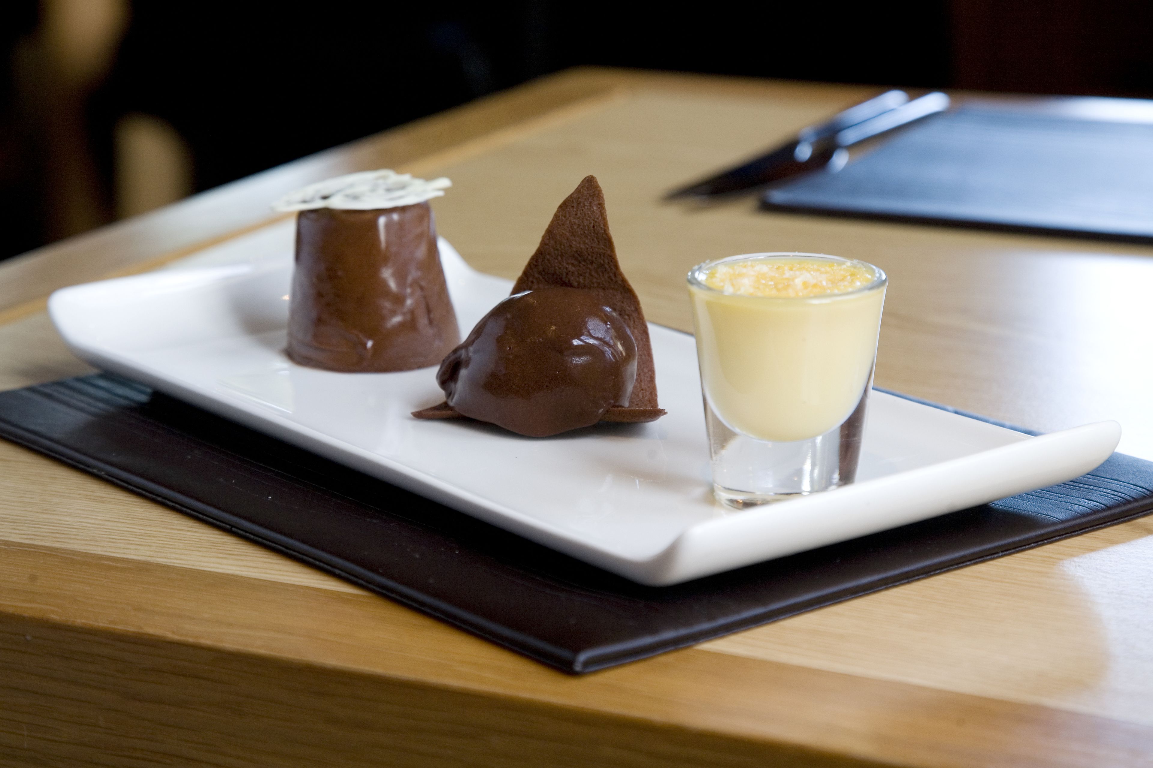 A dessert plate with three chocolate confections and a glass of a creamy beverage on a wooden table.