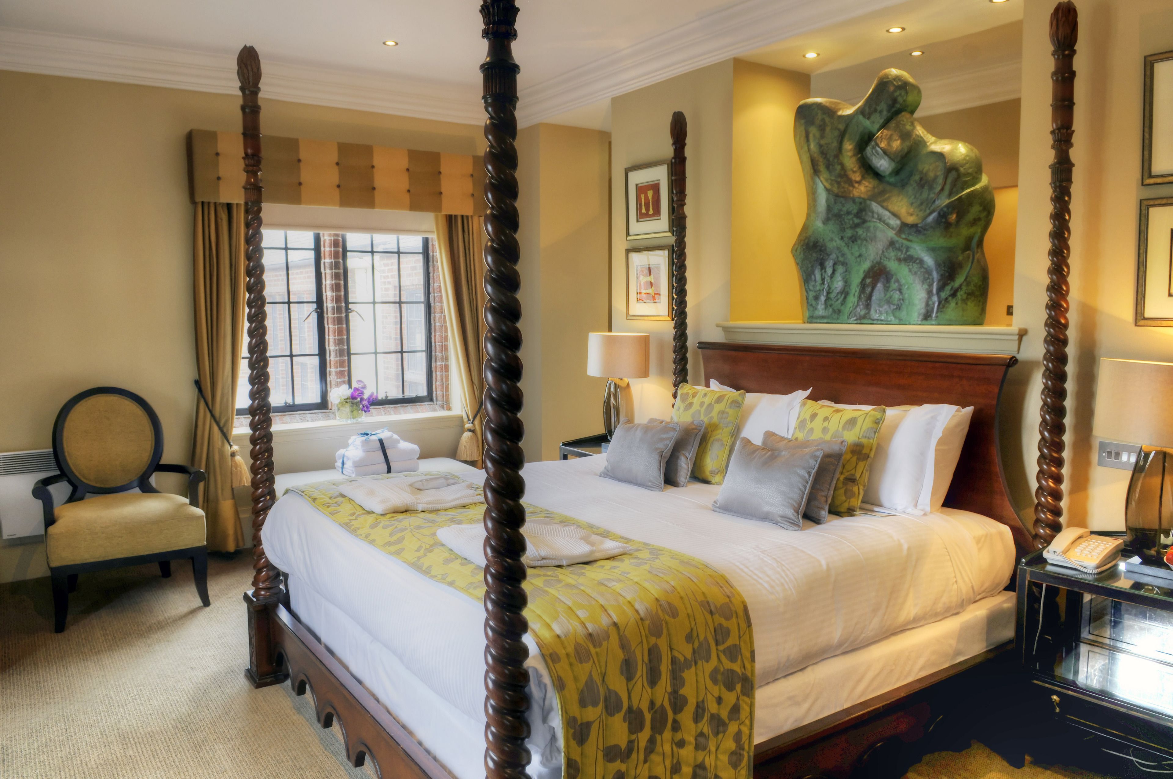 A four-poster bed with a canopy, a chair, a nightstand with a lamp, and a painting on the wall.