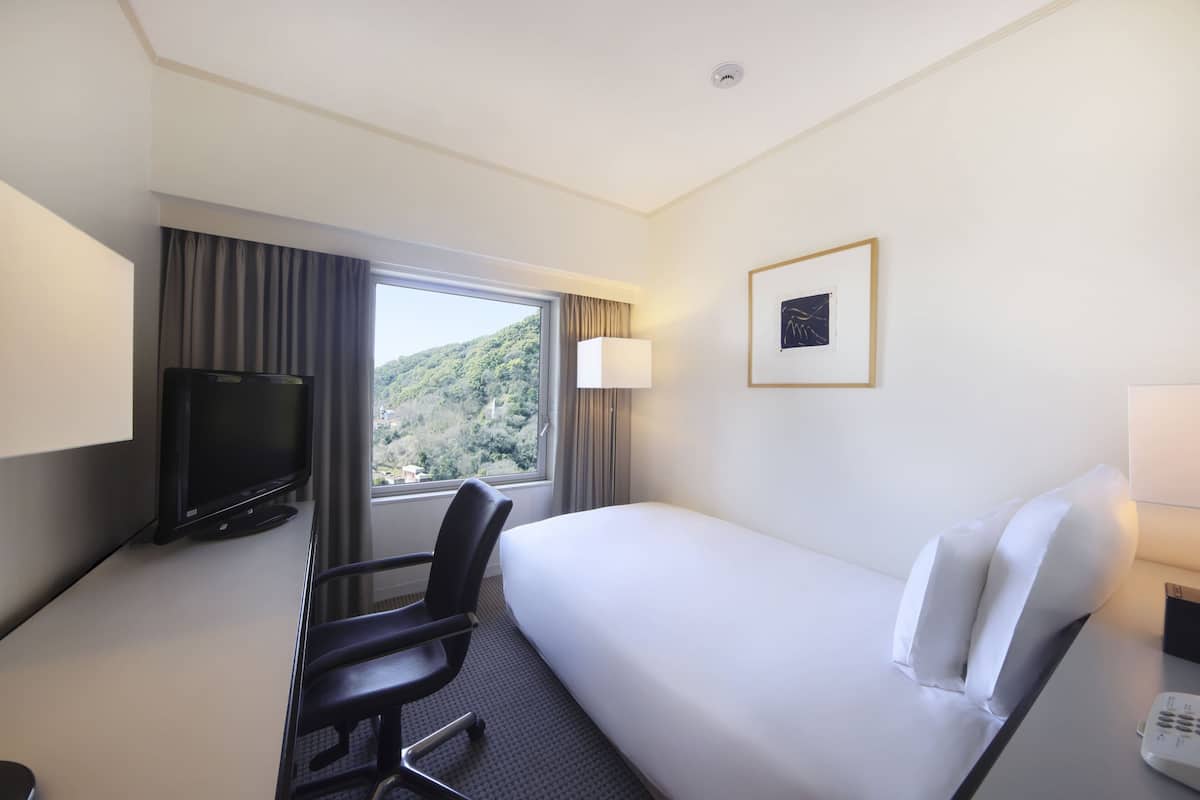 A hotel room with a bed, a desk with a TV, a chair, a window with curtains, a framed picture on the wall, and a bedside table with a lamp.