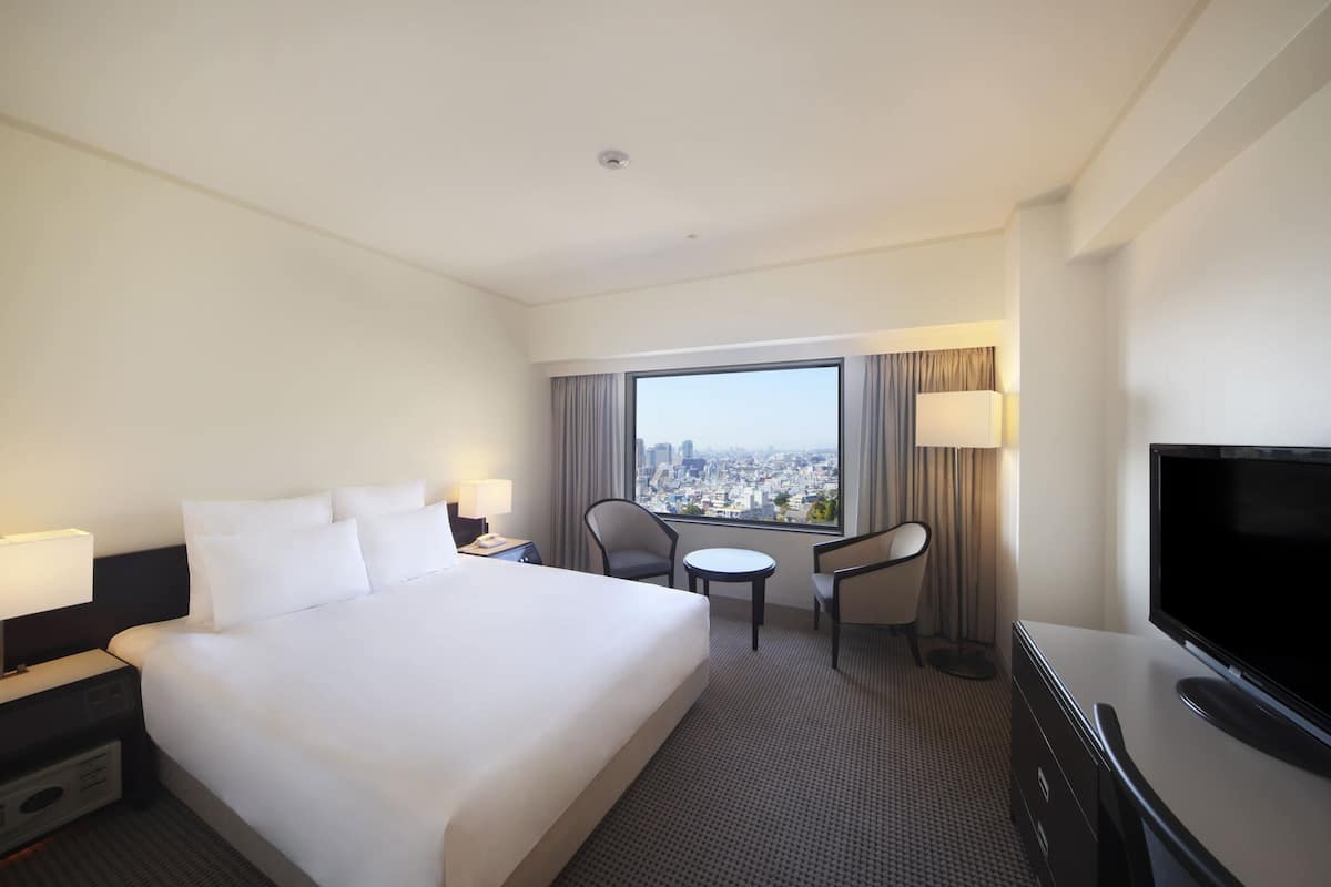 A hotel room with a large bed, a desk with a TV, a chair, and a view of the city through a large window.