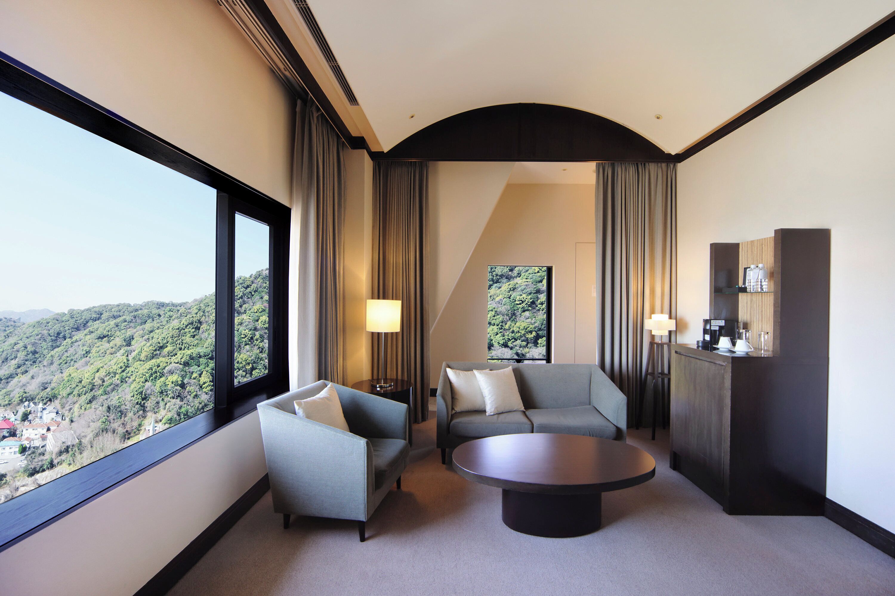 A hotel room with a large window, two sofas, a coffee table, and a minibar.