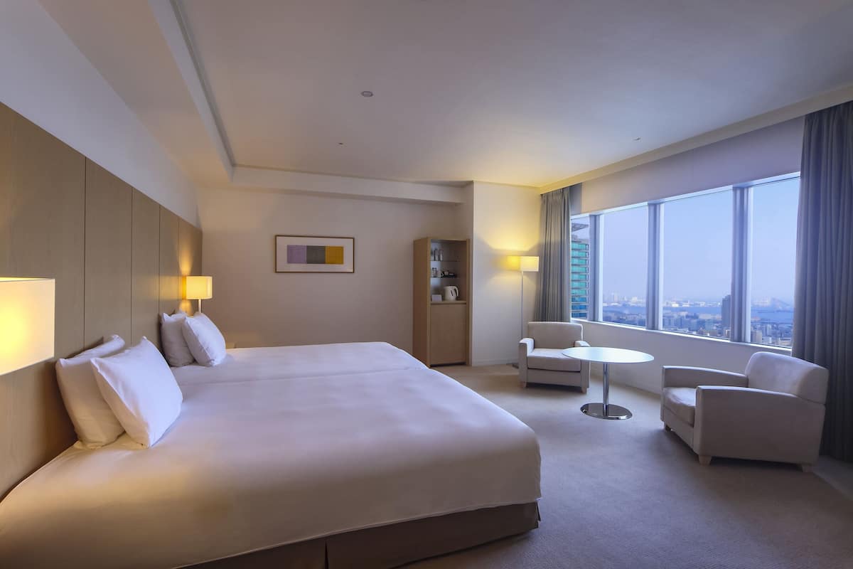 A hotel room with a large bed, two armchairs, a small table, a bookshelf, and a view of the city through large windows.